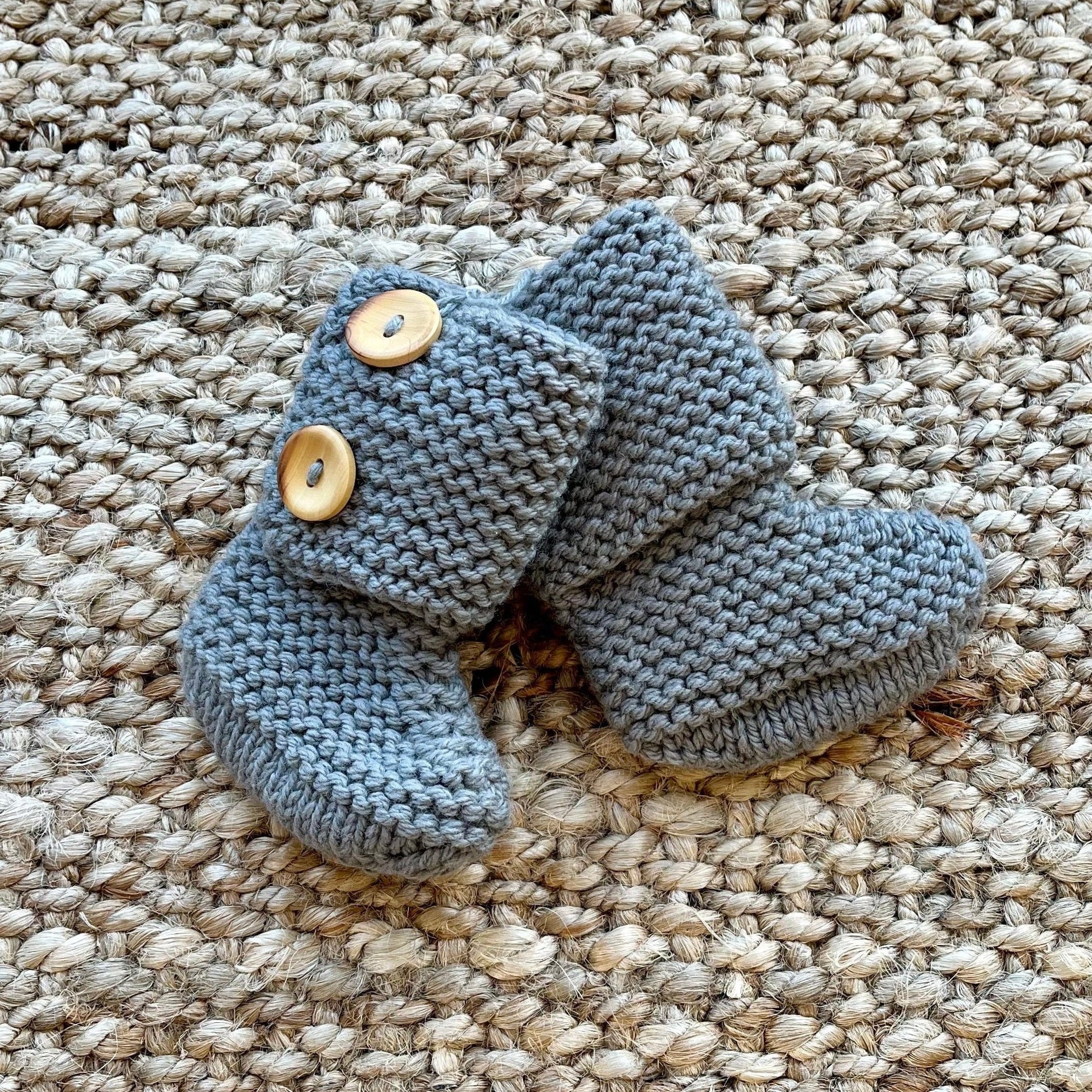 A pair of handmade gray knitted baby booties with two wooden buttons, displayed on a textured beige woven rug.