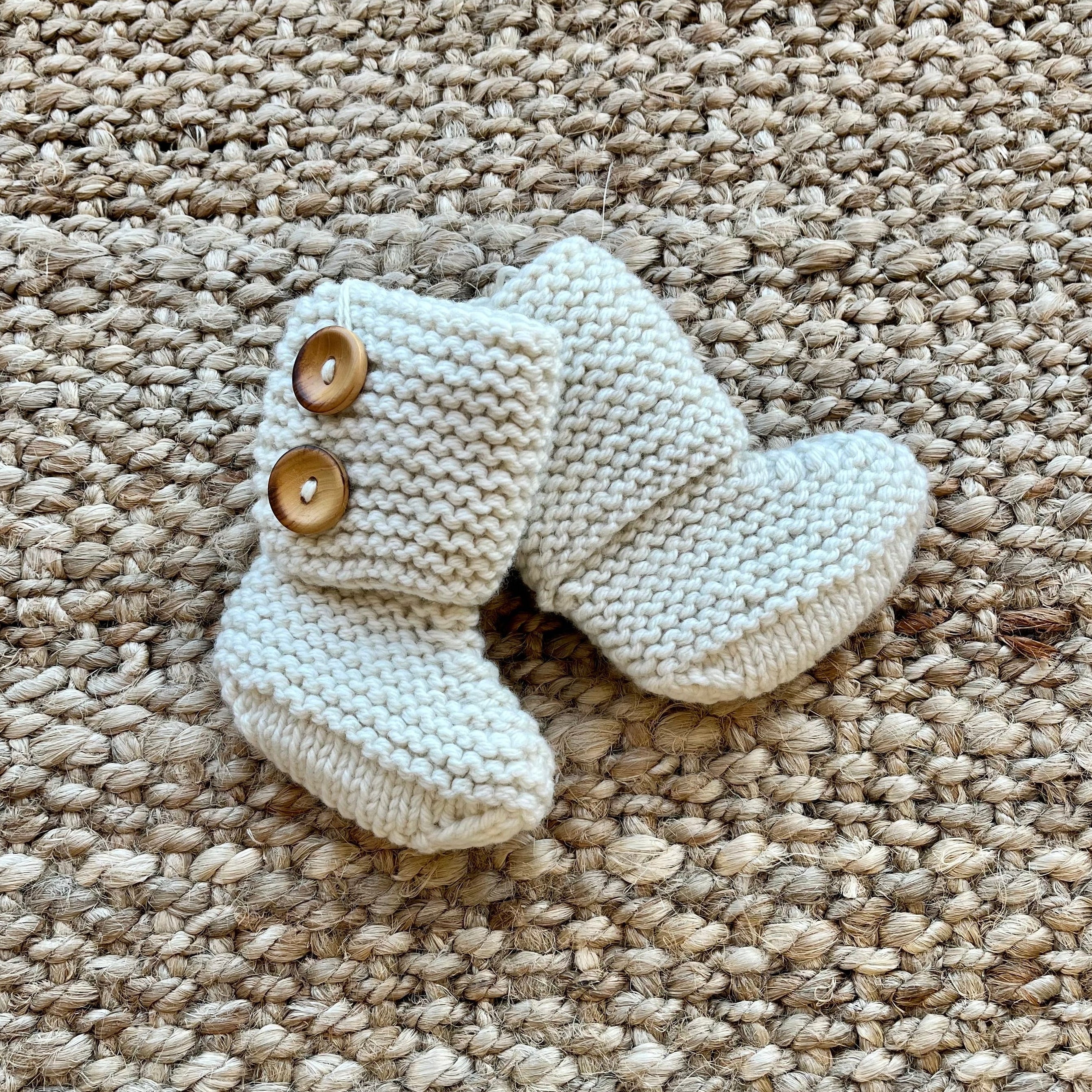 A pair of cream-colored hand-knit baby booties with two wooden buttons, displayed on a textured beige woven rug.