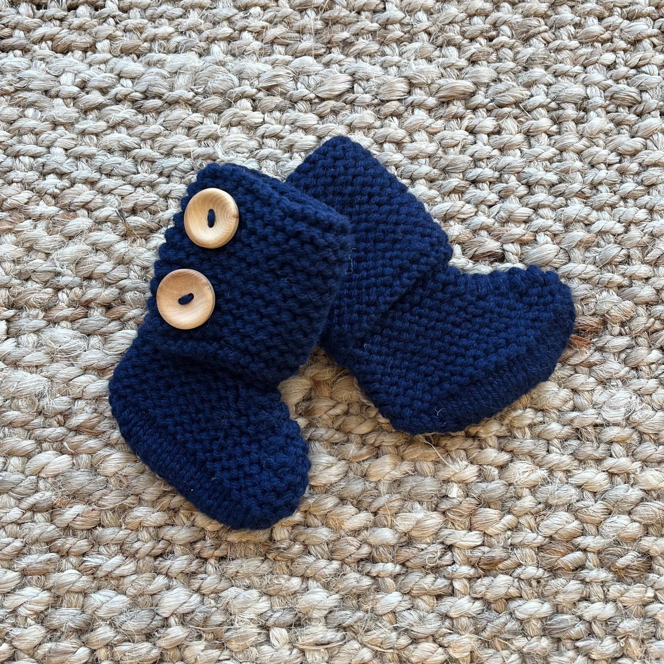 A pair of navy blue hand-knitted baby booties with two light wood buttons, displayed on a textured beige jute rug.