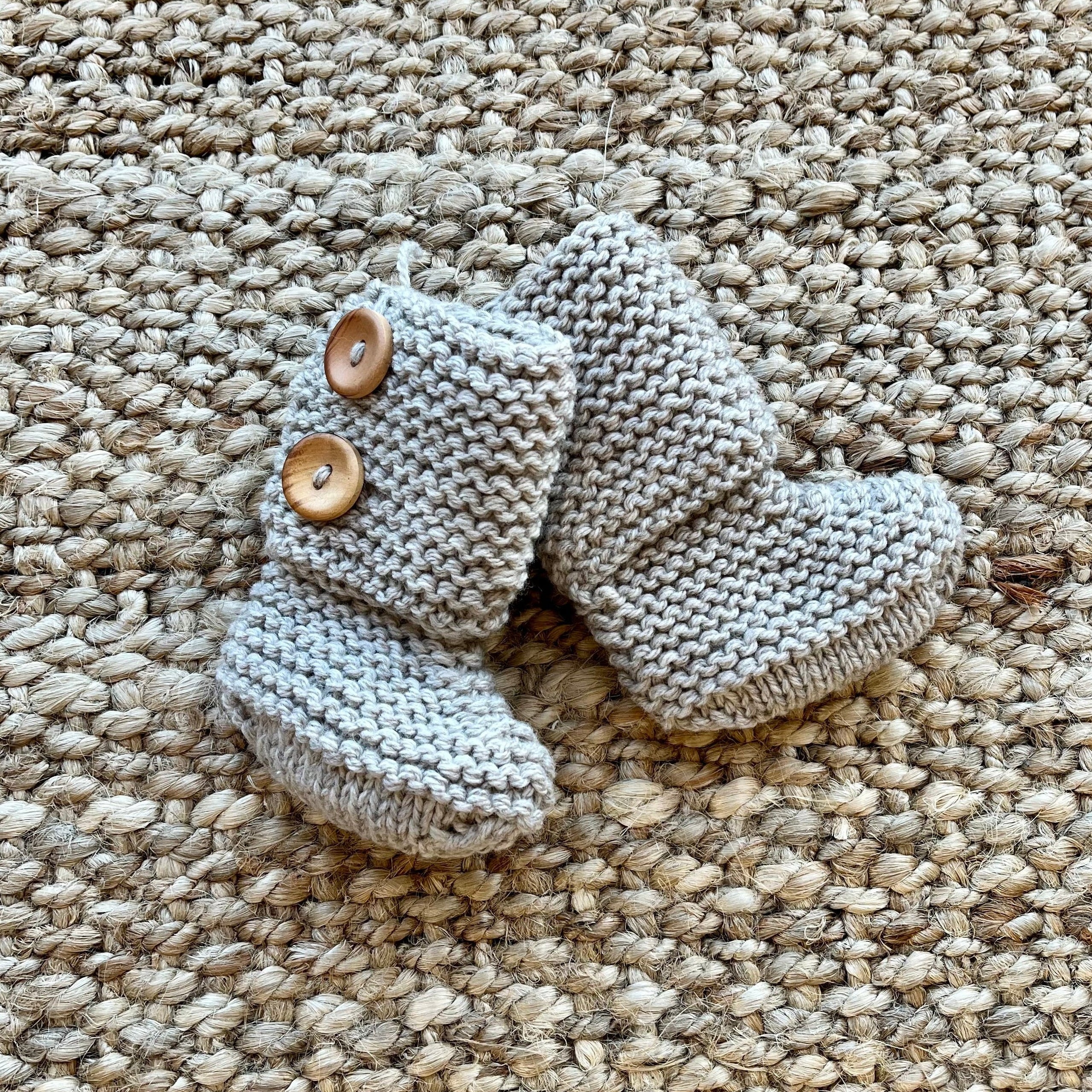 A pair of oatmeal hand-knit baby booties with two wooden buttons, laid flat on a textured beige woven rug.