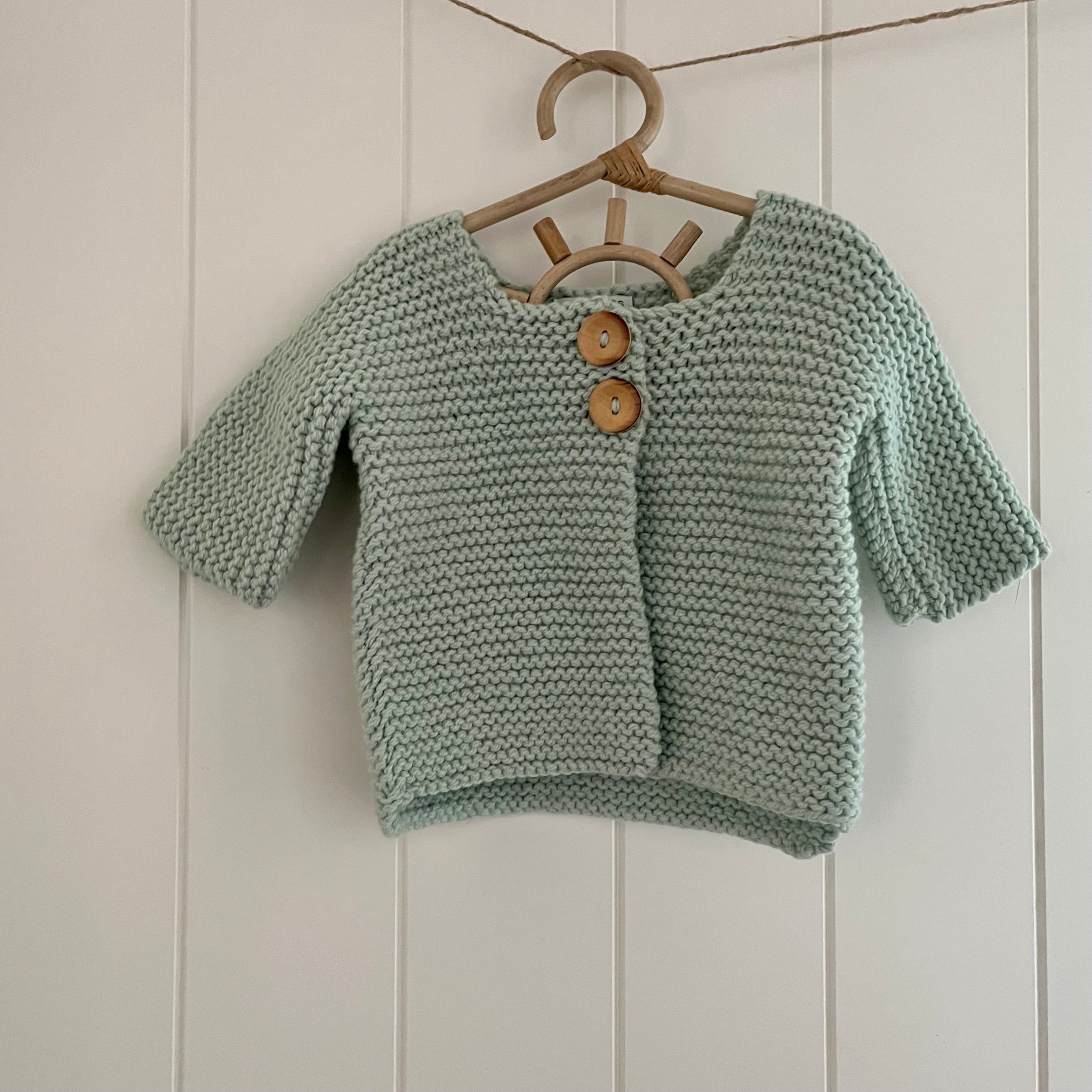 Knitted green baby cardigan with wooden buttons on a hanger against a white paneled wall.