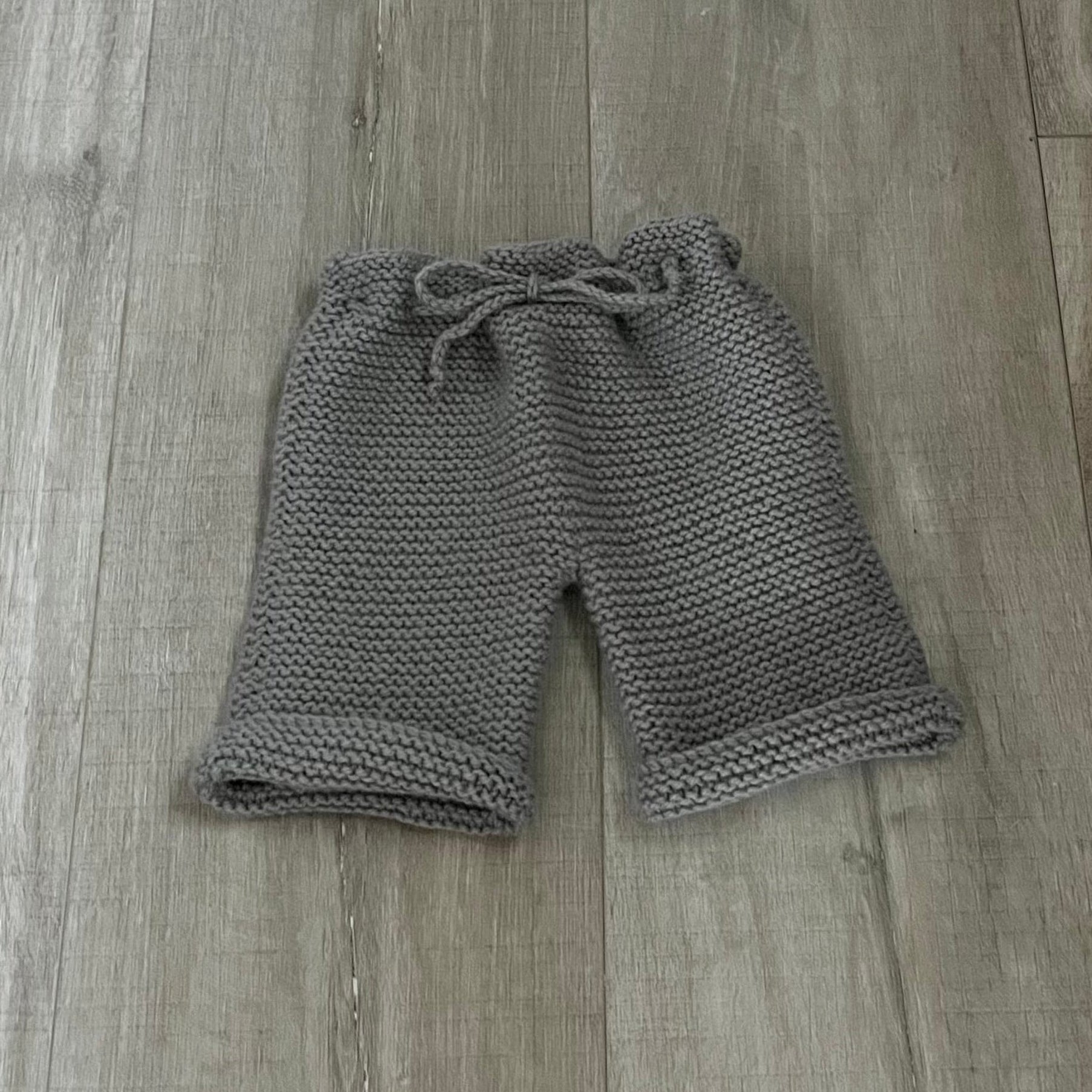 A pair of gray chunky-knit baby pants with a drawstring waist, laid flat on a light brown wooden floor.