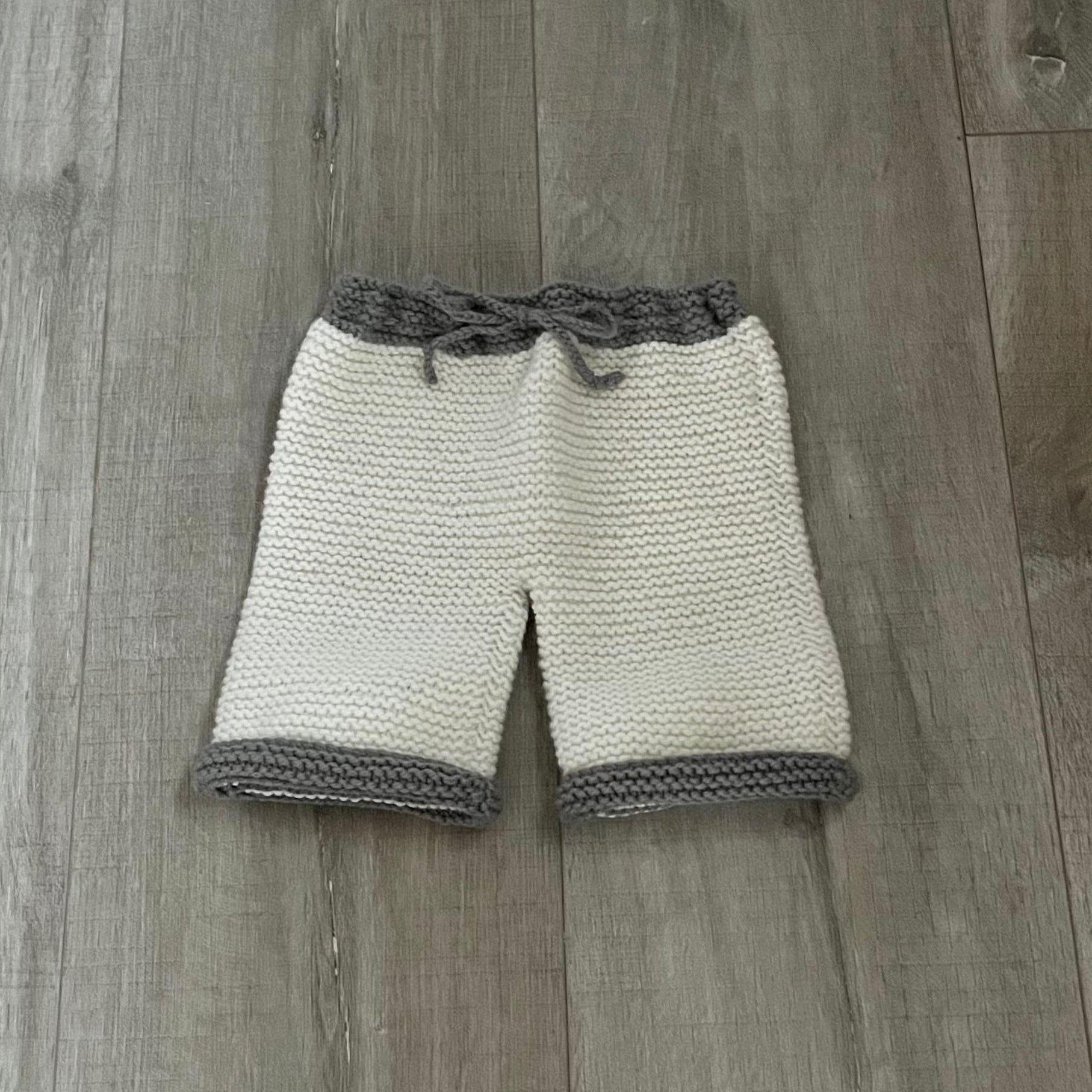 Hand-knit baby pants in cream and gray, laid flat on a gray wood plank floor. The shorts feature a cream knit body, gray knit trim at the cuffs and waistband, and a gray knotted drawstring at the waist.