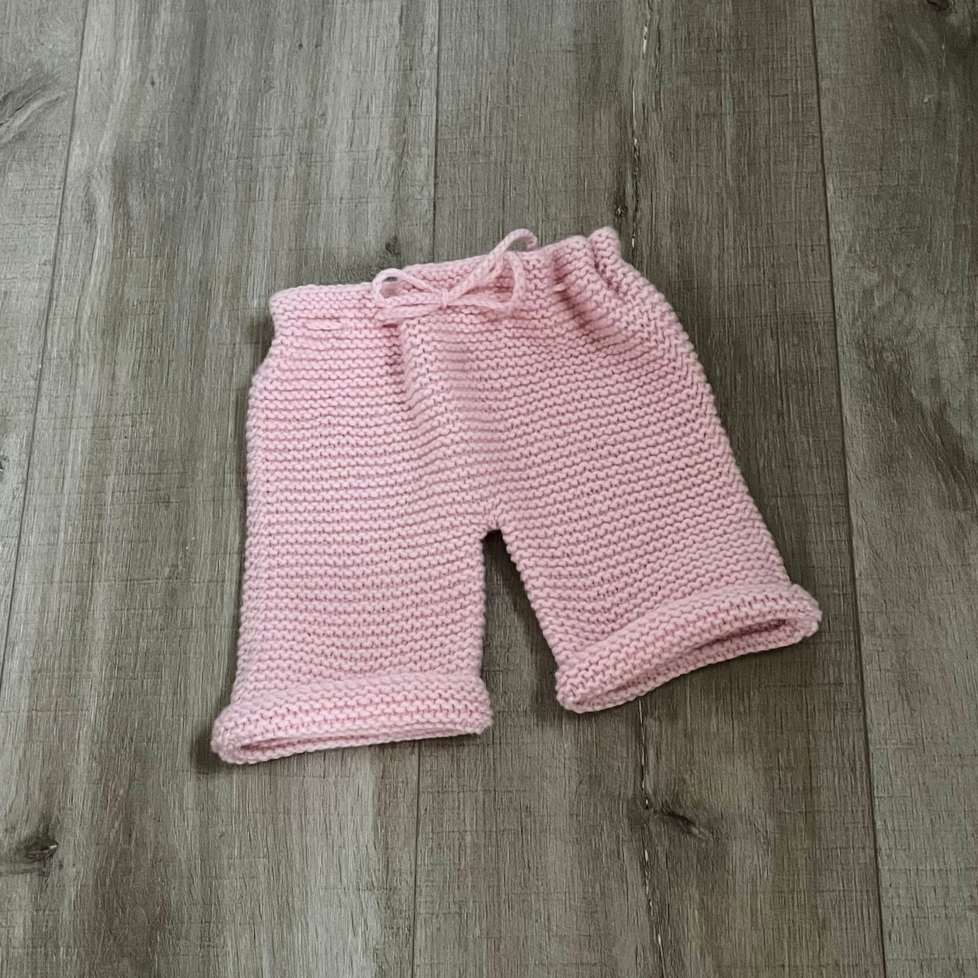 A pair of pink hand-knit baby pants with a drawstring waist, laid flat on a gray-brown wooden floor.