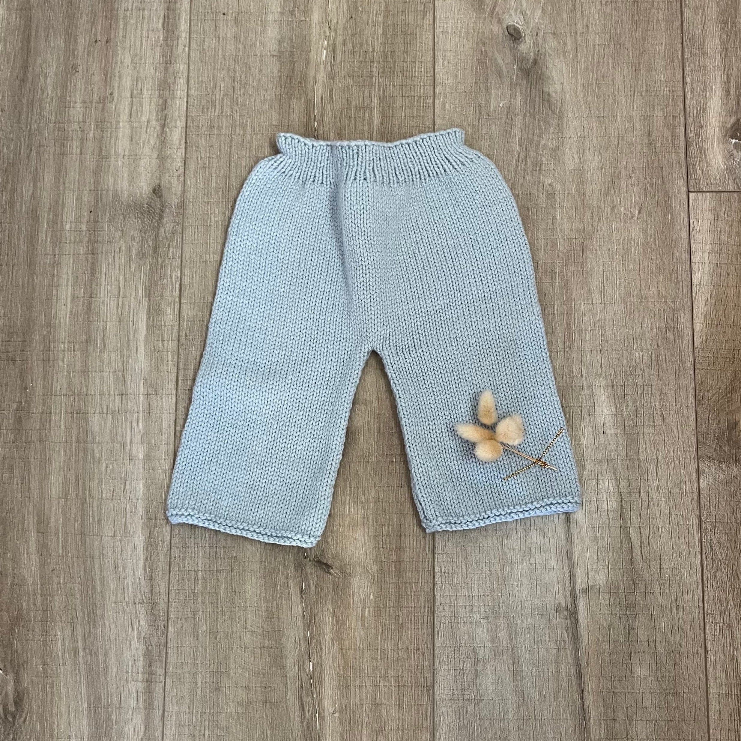 A pair of light blue hand-knit baby pants with an elastic waistband, laid flat on a brown wooden floor with a small dried flower prop near the hem.