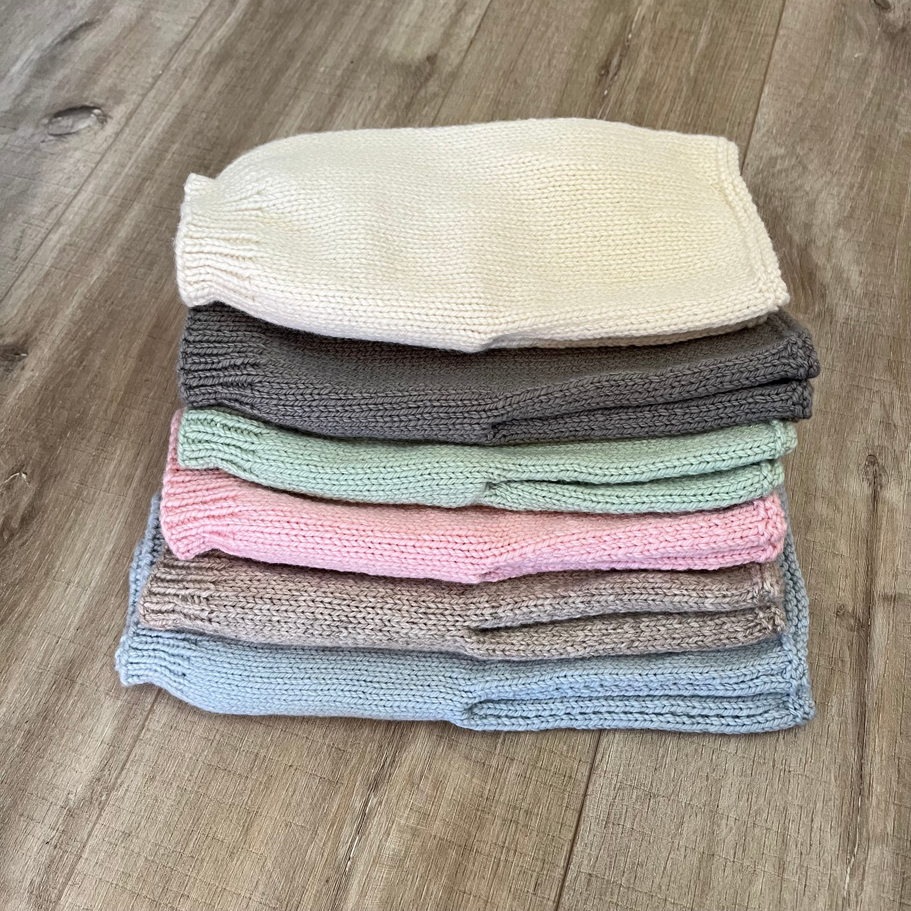 A stack of five knitted baby pants in different pastel and neutral colors, including cream, dark gray, mint green, pink, taupe, and light blue, arranged on a wooden floor.