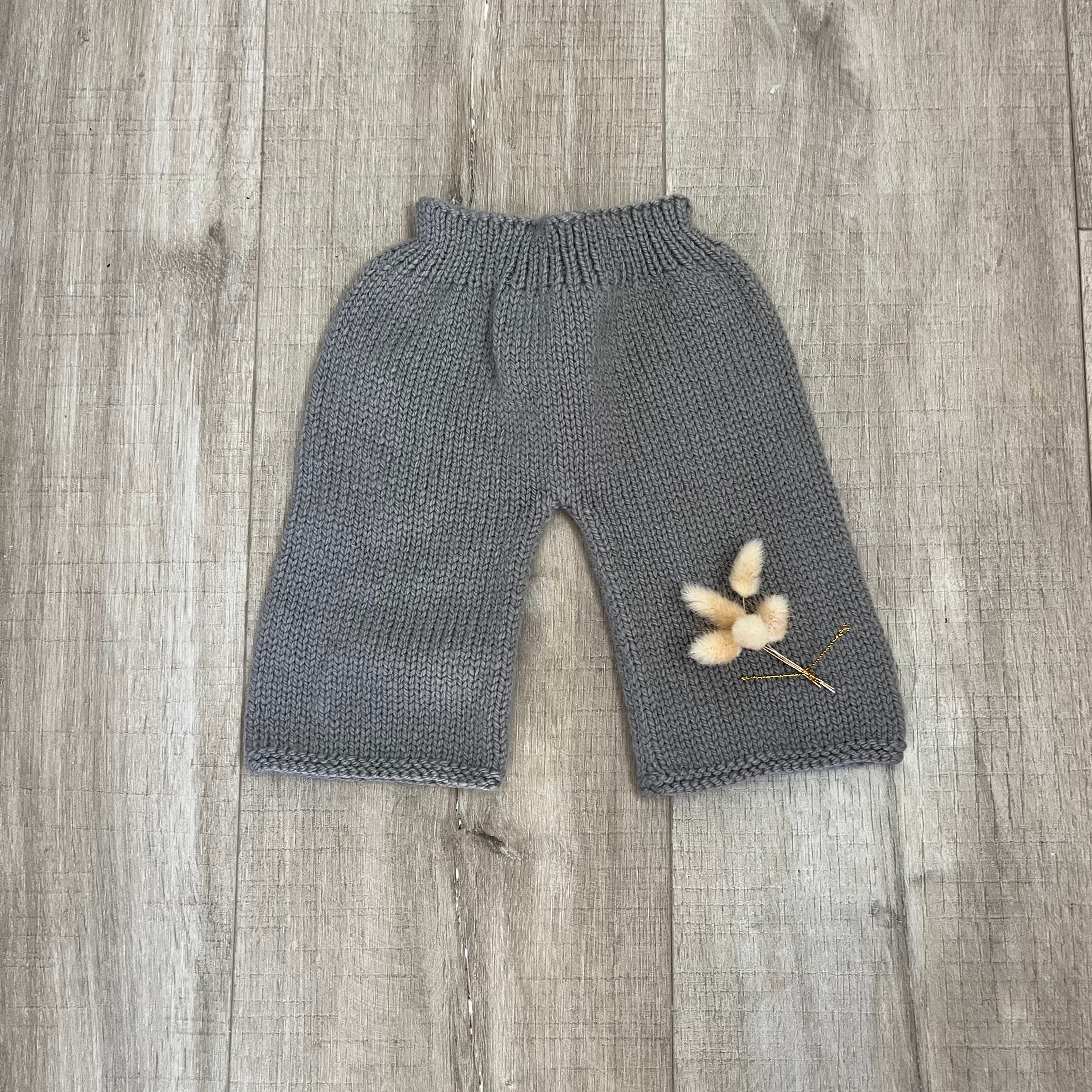 A pair of gray knit baby pants with an elastic waistband, laid flat on a gray wood plank floor with a small dried flower prop.