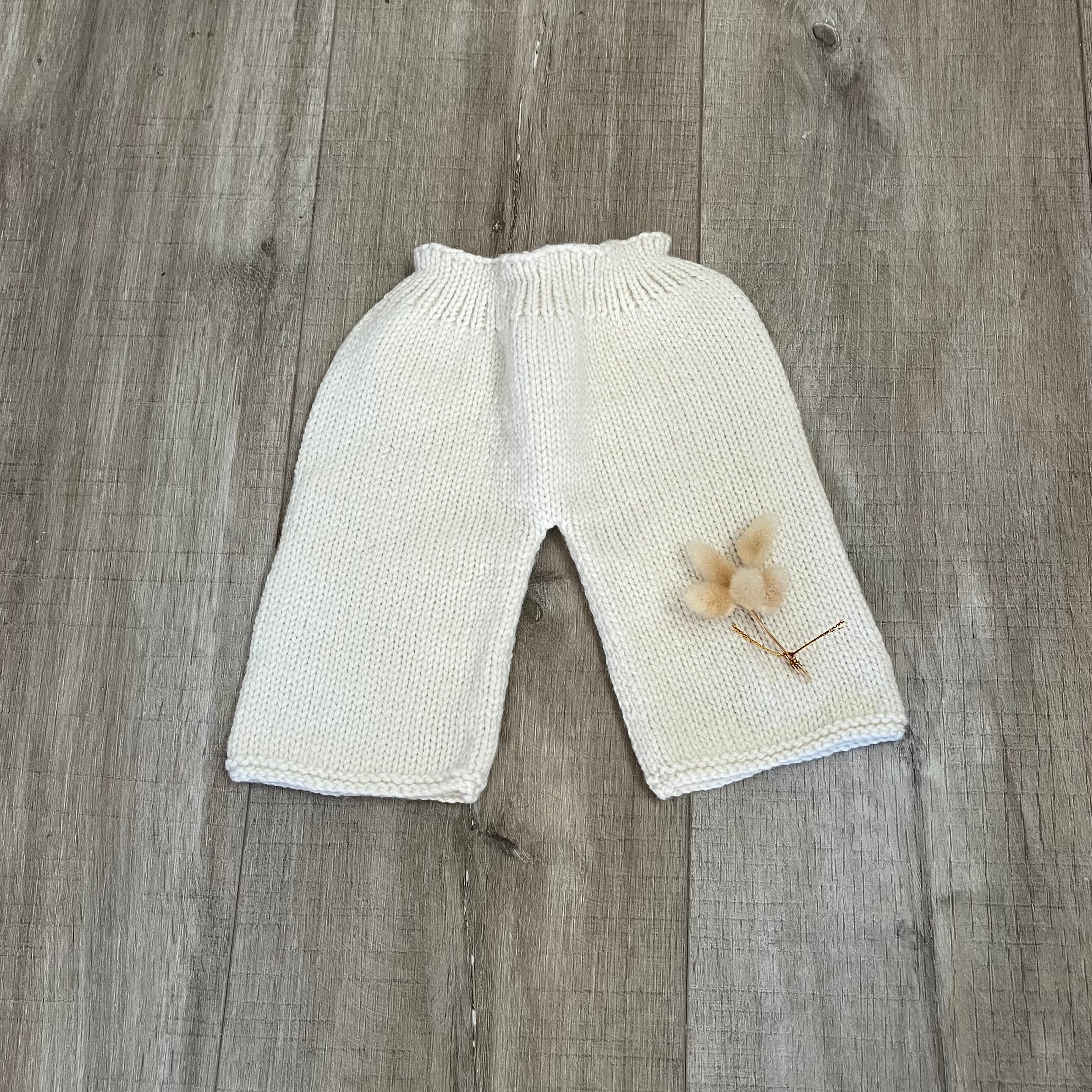 Cream-colored knit baby pants with a small dried flower decoration, laid flat on a gray wood plank floor.
