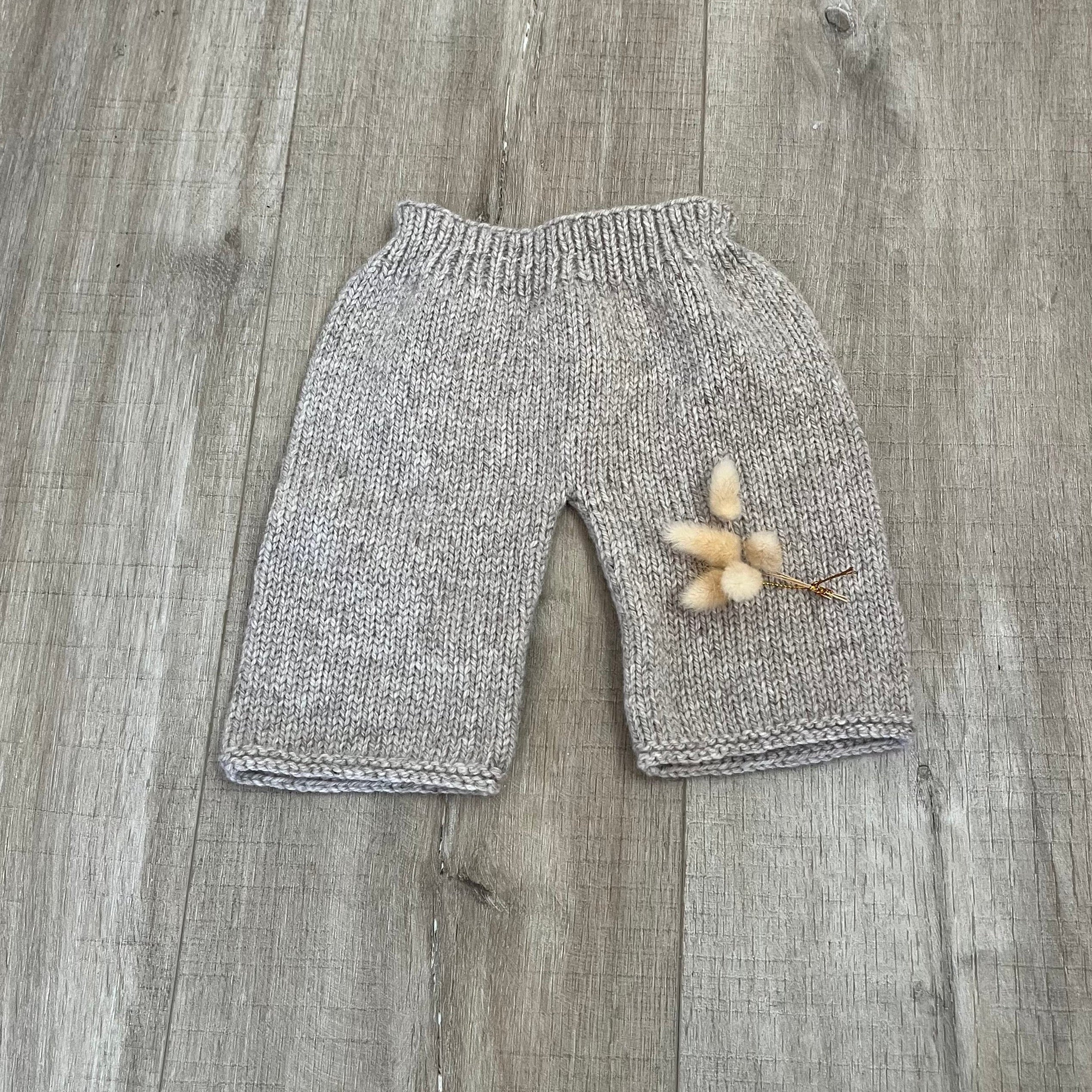 A pair of baby knit pants in a heathered light gray and white yarn, laid flat on a gray wood plank surface with a small dried flower prop.