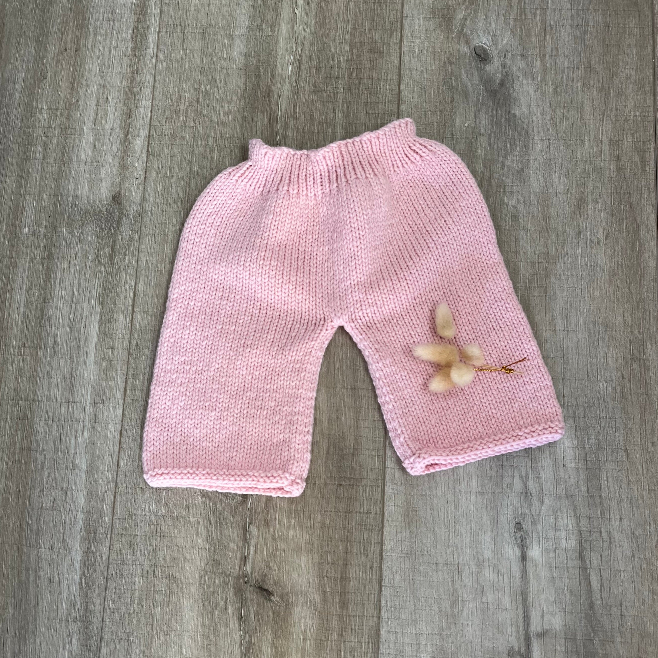 A pair of hand-knit baby pants in light pink, laid flat on a gray wood-plank floor with a small dried flower resting on one leg.