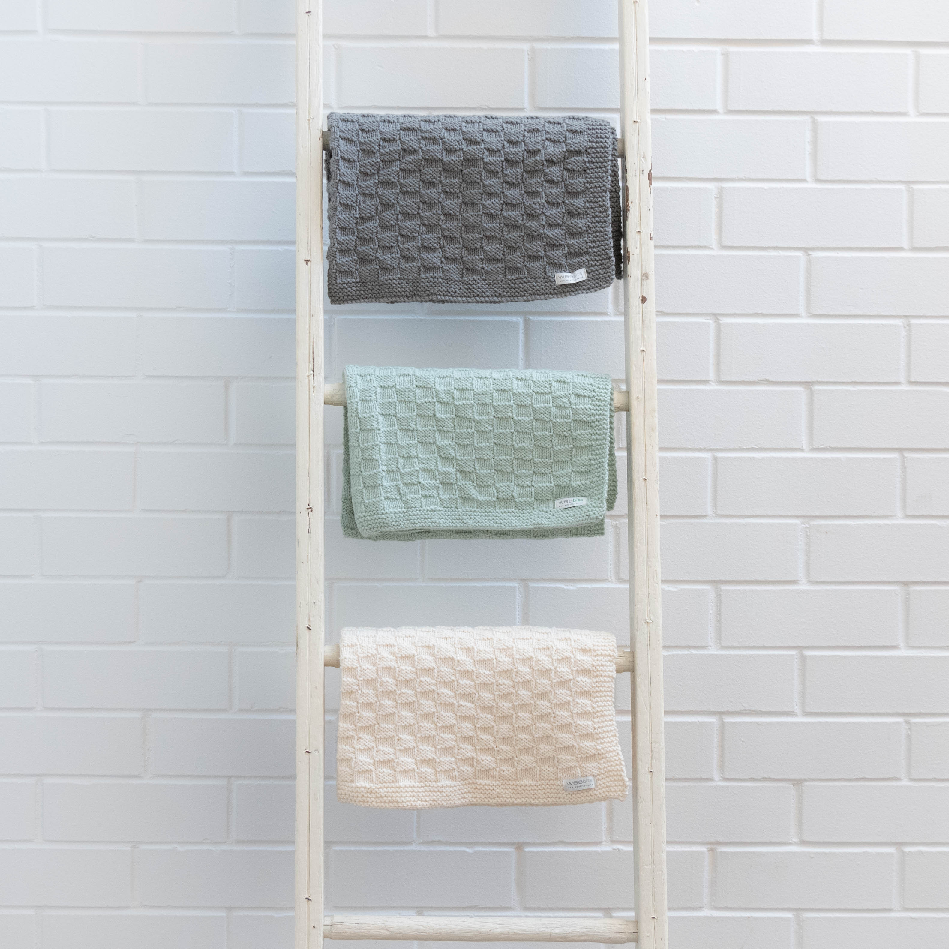 Ladder with three textured trugs in gray, green, and natural against a white tiled wall.