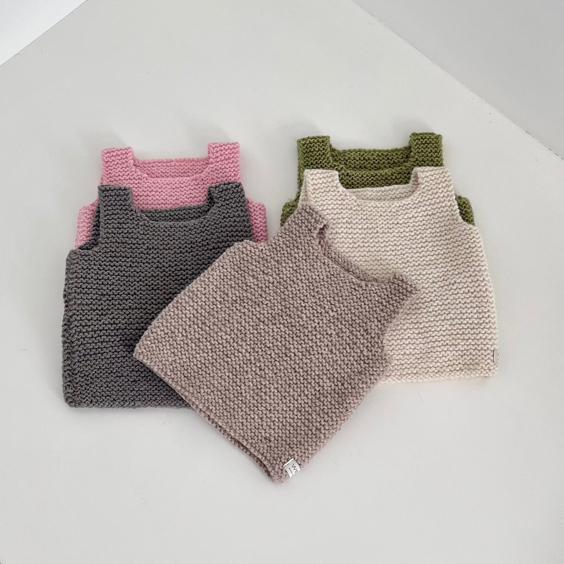 Four knitted sleeveless baby tops in different colors are stacked and arranged on a light gray surface. The tops are in pink, dark gray, taupe, and cream, with the cream top in the foreground.