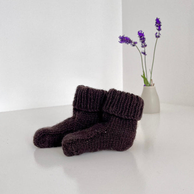 Pair of dark brown knitted baby socks on a white surface with a vase of purple flowers in the background.