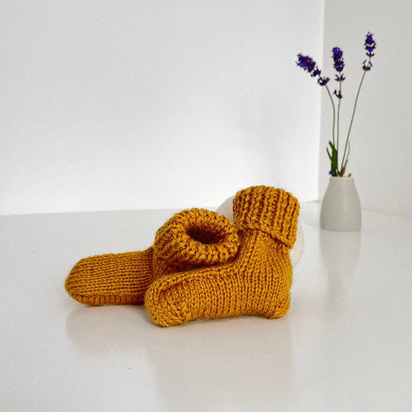 Pair of mustard knitted socks on a white surface with a small vase of lavender in the background.