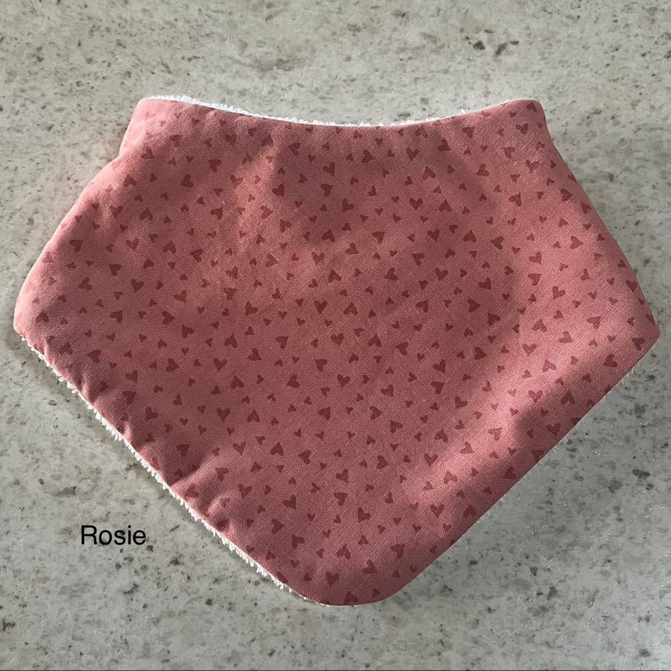 Pink dribble bib with heart pattern on a concrete surface, branded 'Rosie'.