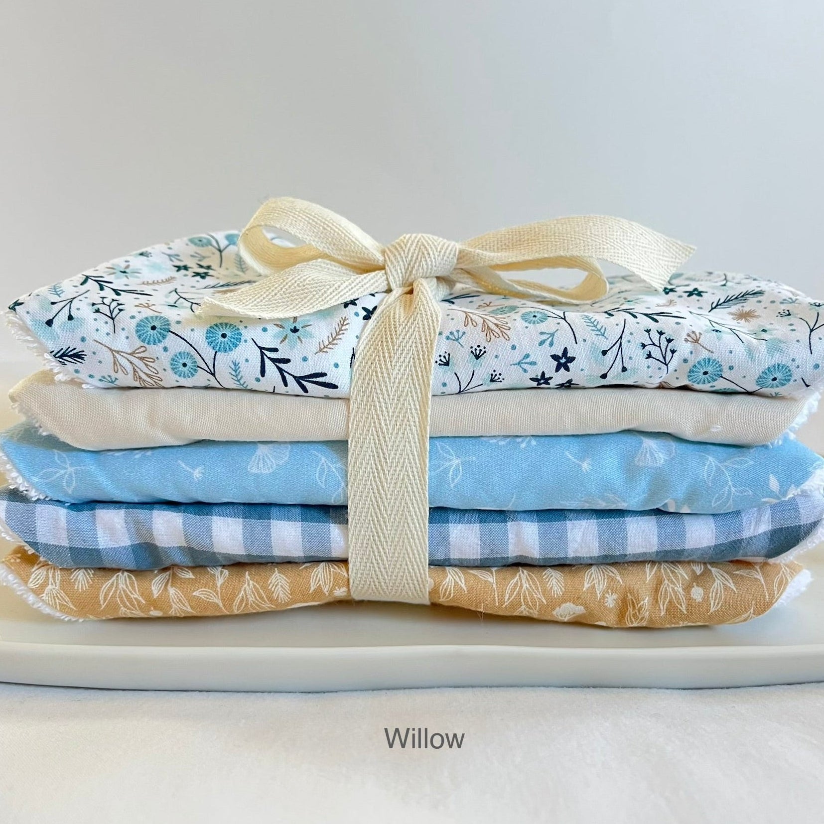 A stack of five folded fabric dribble bibs tied with a cream ribbon, displayed on a white surface. The fabrics feature floral, gingham, and botanical prints in light blue, beige, and white.