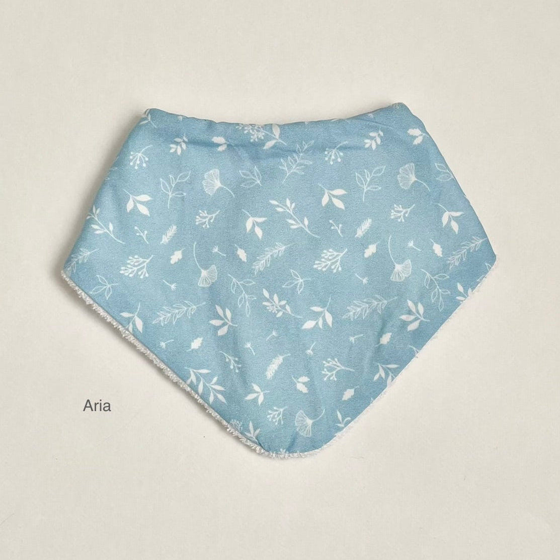 A light blue triangular bandana bib with a white floral and leaf pattern, laid flat on a neutral cream background. The name 'Aria' is printed in black text to the left of the bib.