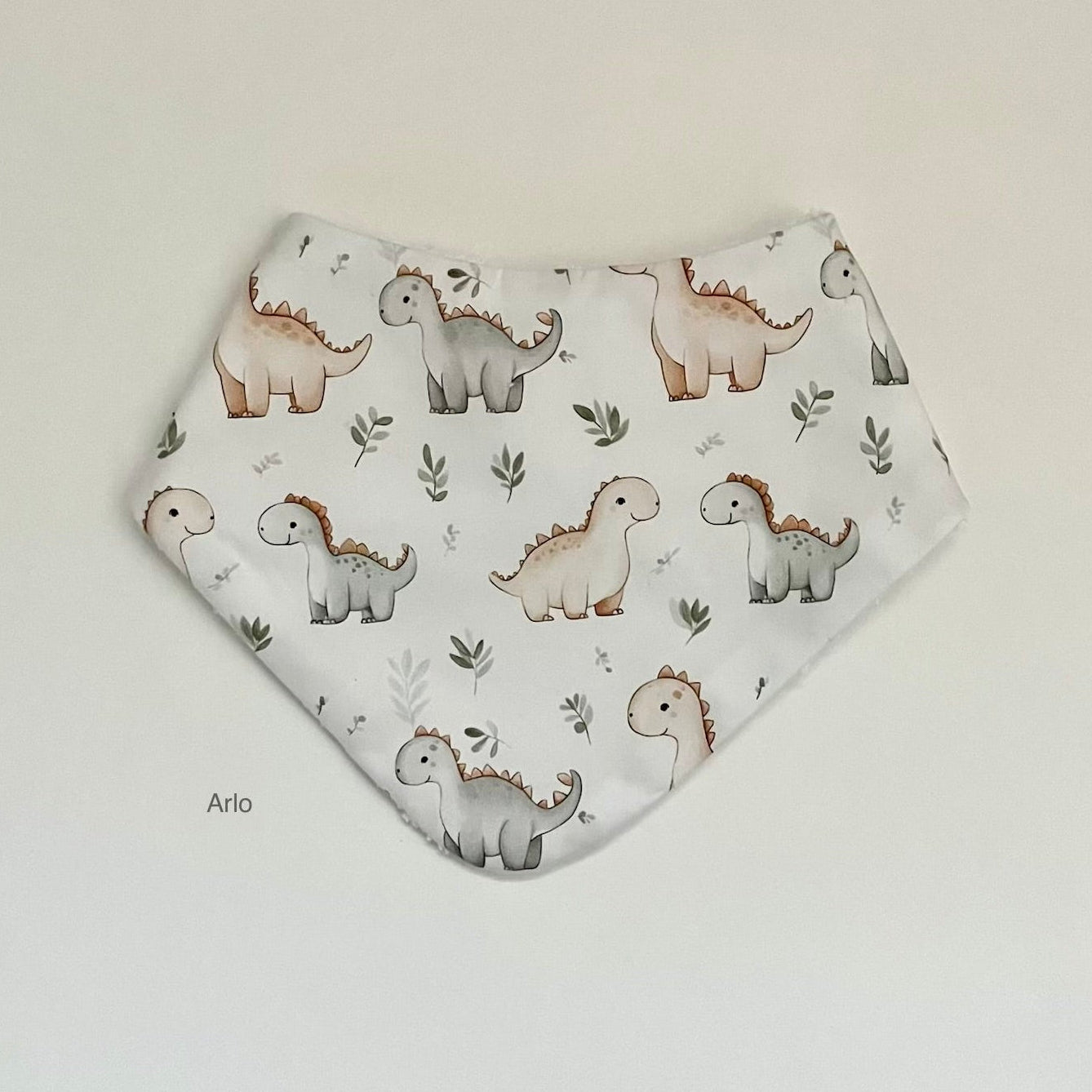 A triangular bandana bib with a white background and a pattern of cute cartoon dinosaurs in brown, gray, and beige, accented with small green leaves. The bib is laid flat on a neutral light gray background.