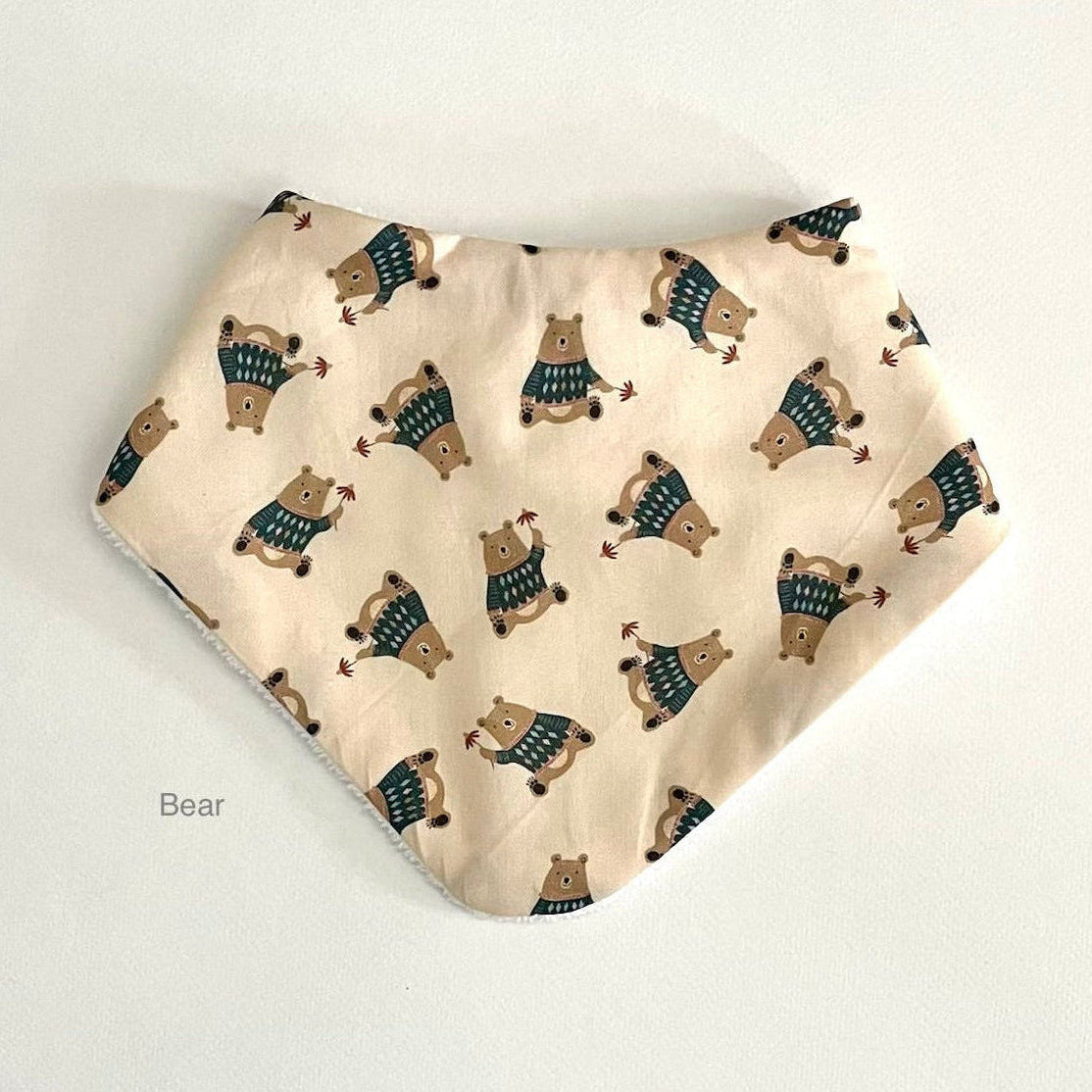 A triangular bandana bib with a cream background and an all-over print of cartoon bears wearing blue and green sweaters, shown on a plain light background.