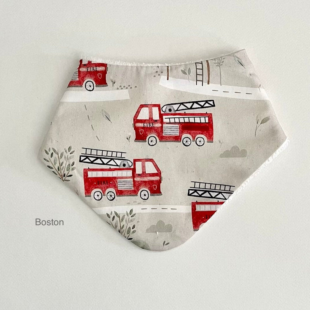A triangular baby bandana bib with a red fire truck pattern on a light beige background, displayed flat against a white background with the text 'Boston' printed on the bottom left.