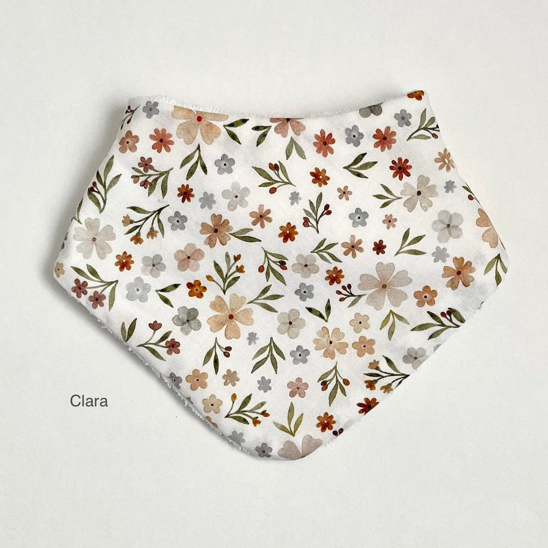 A white fabric bib with a delicate floral pattern in muted shades of brown, beige, gray, and green, laid flat on a light off-white background. The name 'Clara' is printed in a small black font in the lower-left corner.