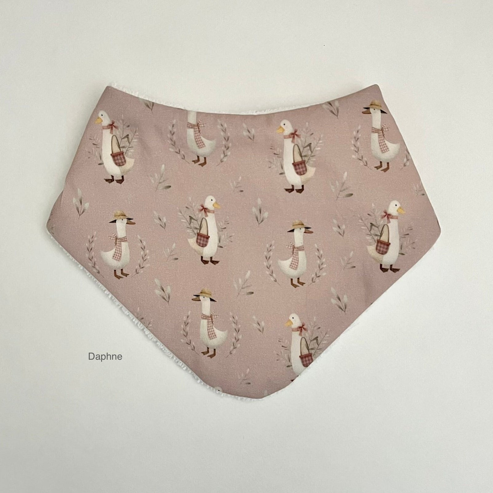 A triangular bandana bib in dusty pink with a repeating pattern of white ducks wearing brown scarves and hats, set against a plain light background.