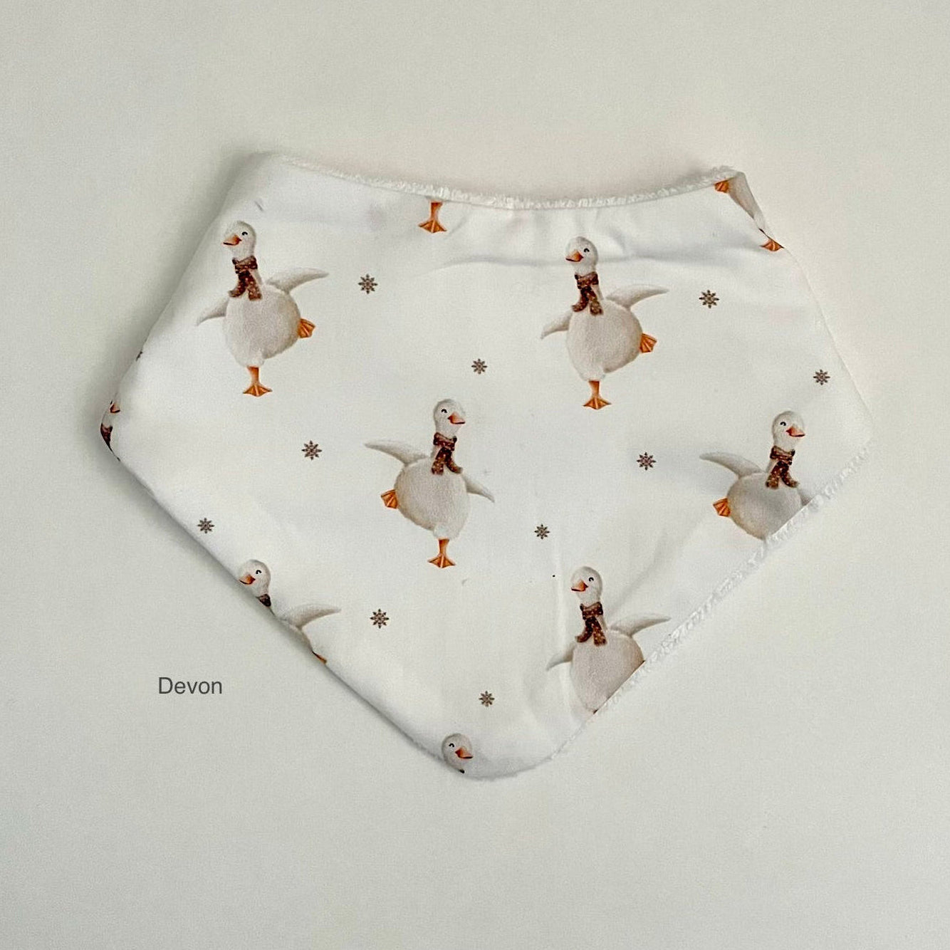 A white reusable cloth bib featuring a repeating pattern of cartoon geese wearing brown scarves and orange beaks, with small gray snowflake motifs. The bib is laid flat on a light gray background, with the brand name 'Devon' printed in a small gray font in the lower-left corner.