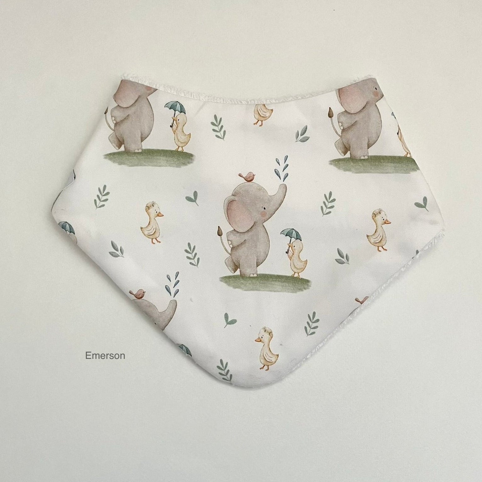 A white bandana bib with a watercolor print of elephants, ducks, and small plants, shown on a light gray background. The brand name 'Emerson' is printed in a small black font on the lower left.