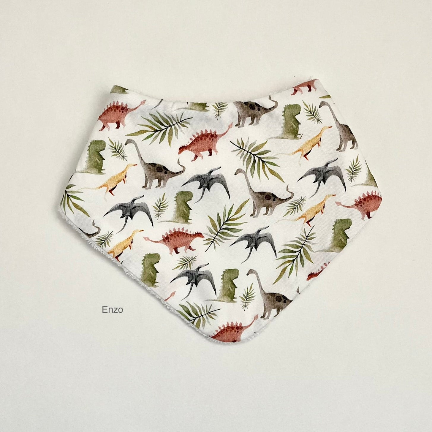 A white triangular bandana bib featuring an all-over print of colorful dinosaurs and palm leaves, laid flat on a light beige background. The brand name 'Enzo' is printed in small black text below the bib.