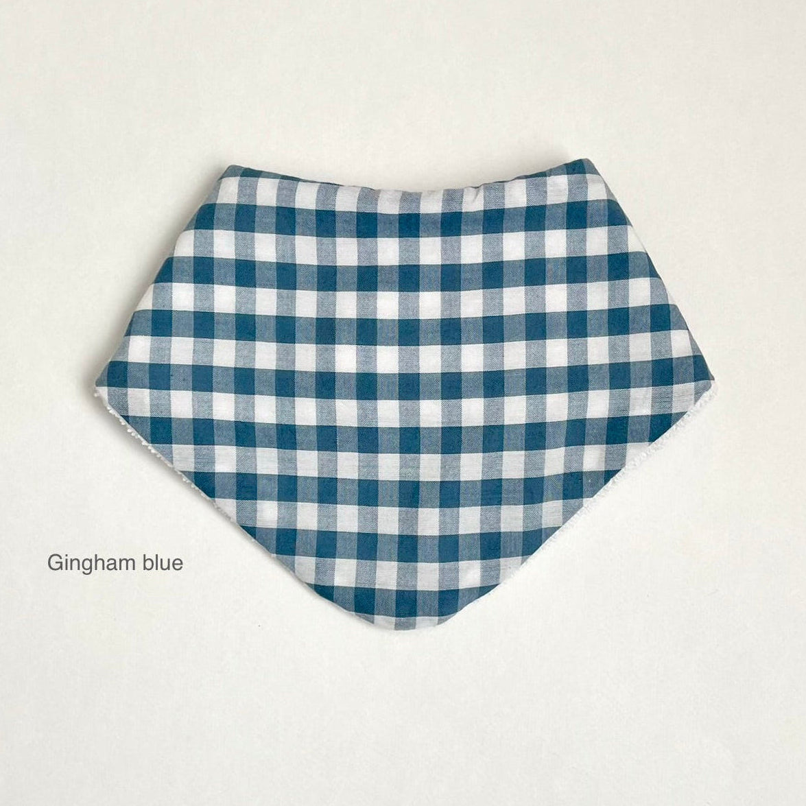 A triangular bandana bib with a blue and white gingham check pattern, displayed flat against a light off-white background with the text 'Gingham blue' printed on the bottom left.
