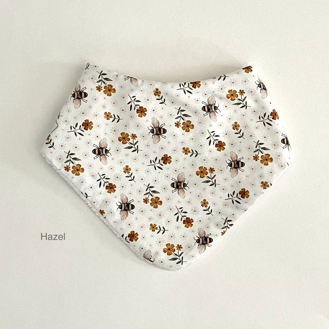 A white bandana-style baby bib with a printed pattern of bumblebees, small brown flowers, and green leaves, laid flat on a light beige background. The text 'Hazel' is printed on the bottom left of the image.