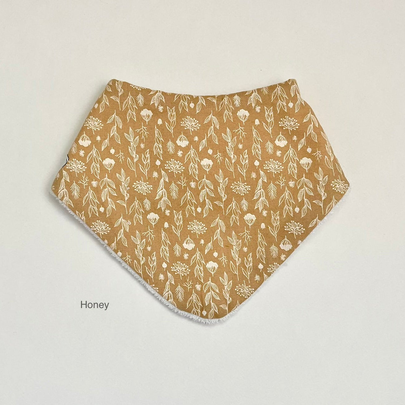 A honey-colored fabric bandana bib with a white floral and leaf pattern, displayed flat against a light off-white background with the text 'Honey' printed below it.