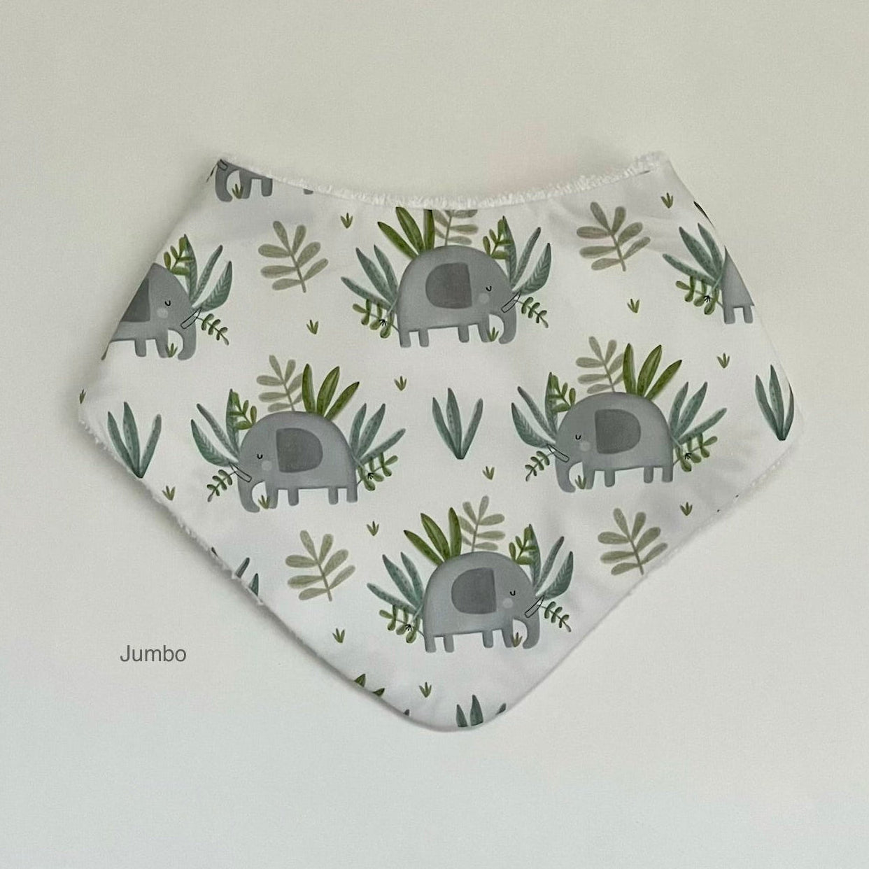 A white triangular bandana bib with a repeating pattern of gray elephants and green leaves, labeled 'Jumbo' on the bottom left, photographed flat against a light background.