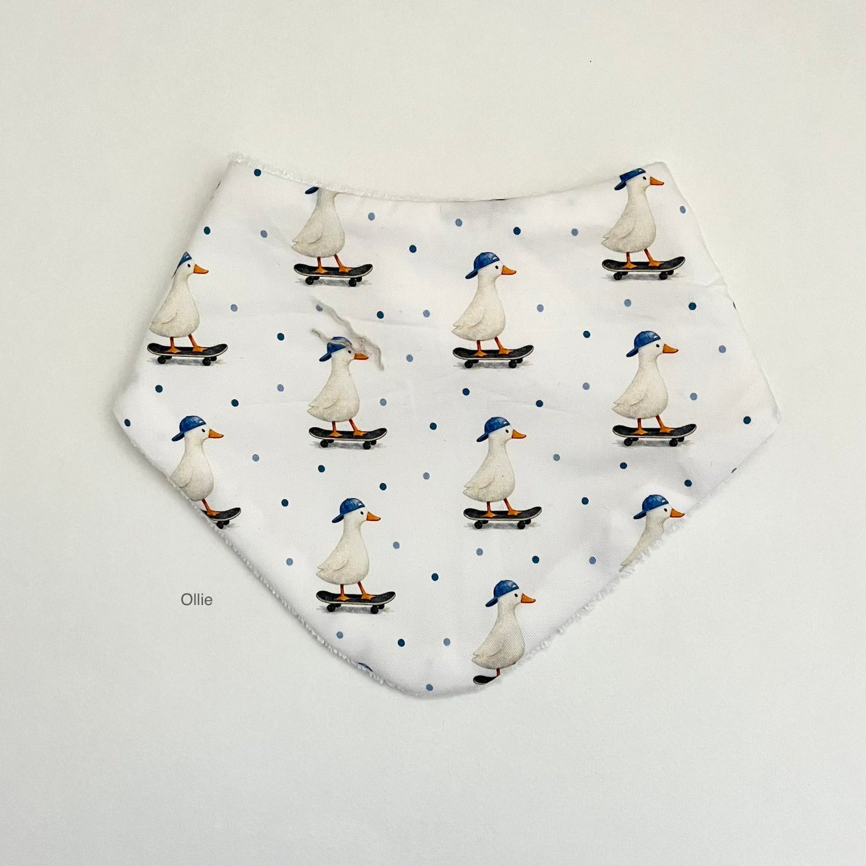 A white bandana bib with a repeating pattern of white ducks wearing blue caps and skateboarding, set against a light gray background.