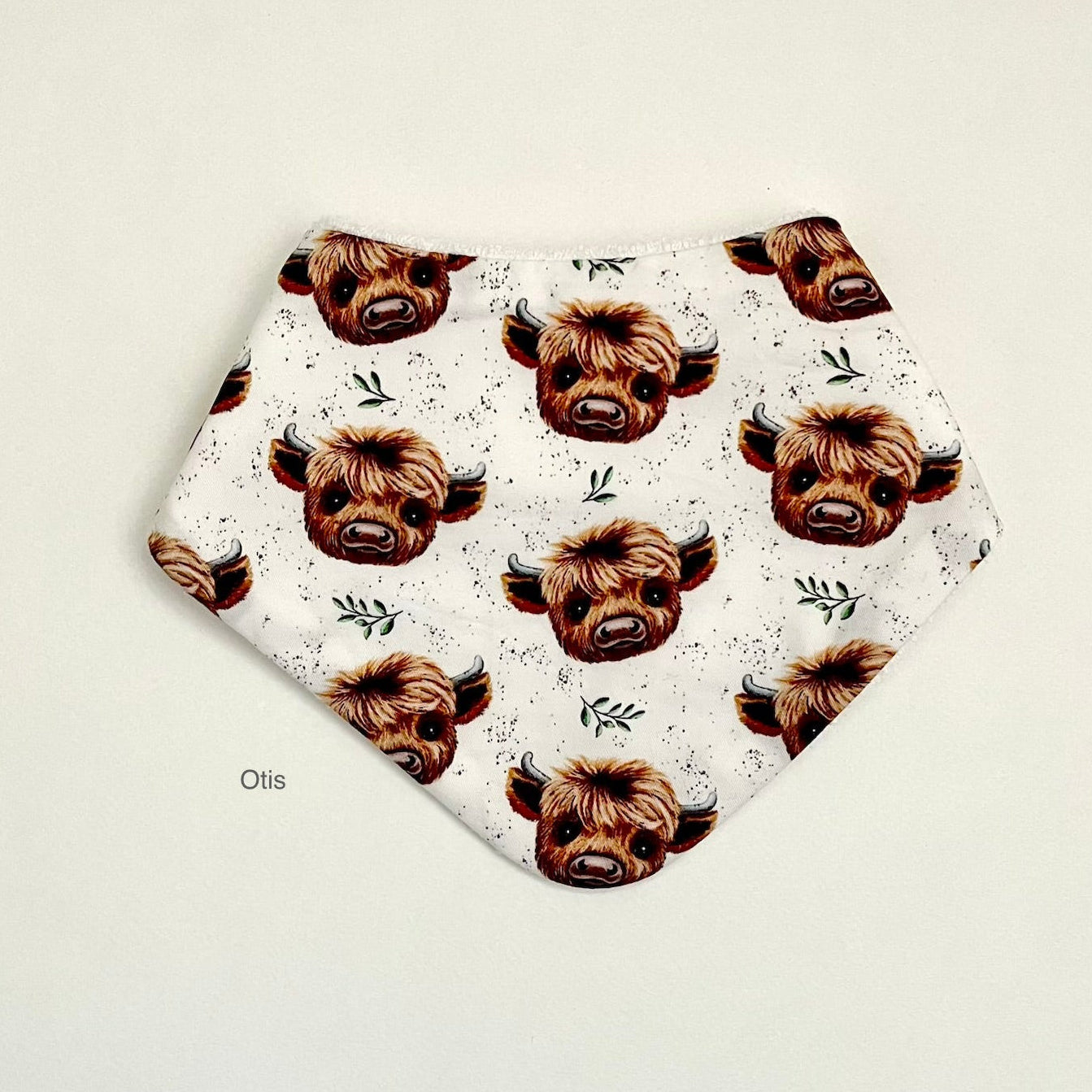 A white hexagonal bandana bib featuring a repeating pattern of brown highland cow faces with small green sprigs and black speckles, displayed flat on a light beige background.