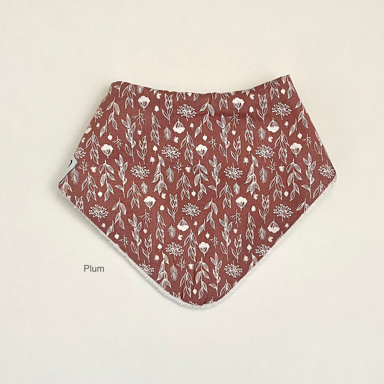 A plum-colored cotton bandana bib with a white floral and leaf pattern, laid flat on a light beige background. The word 'Plum' is printed in small black text near the bottom left corner of the bib.