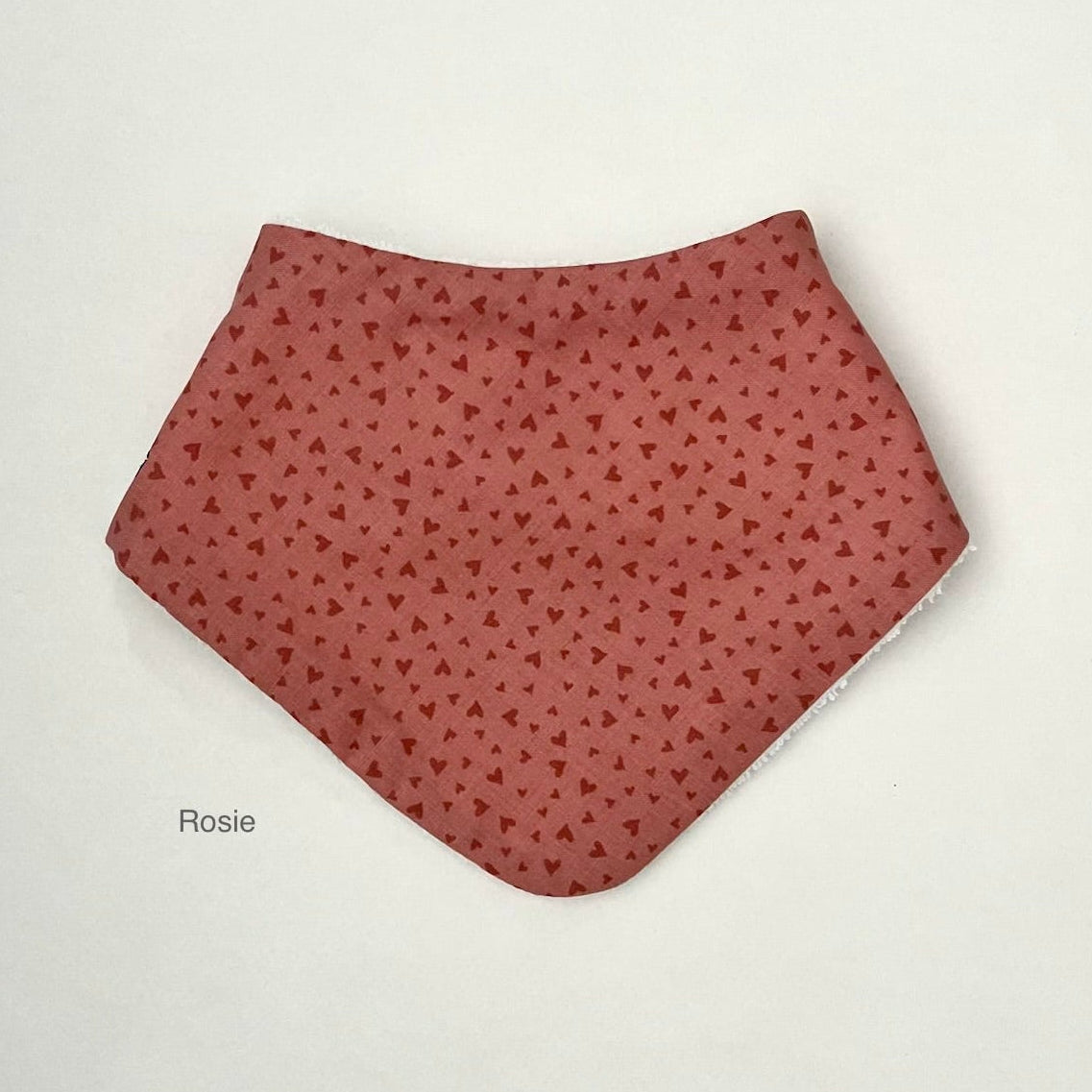 A terracotta red bandana bib with a small repeating dark red heart pattern, displayed flat on a light off-white background with the text 'Rosie' printed below it.