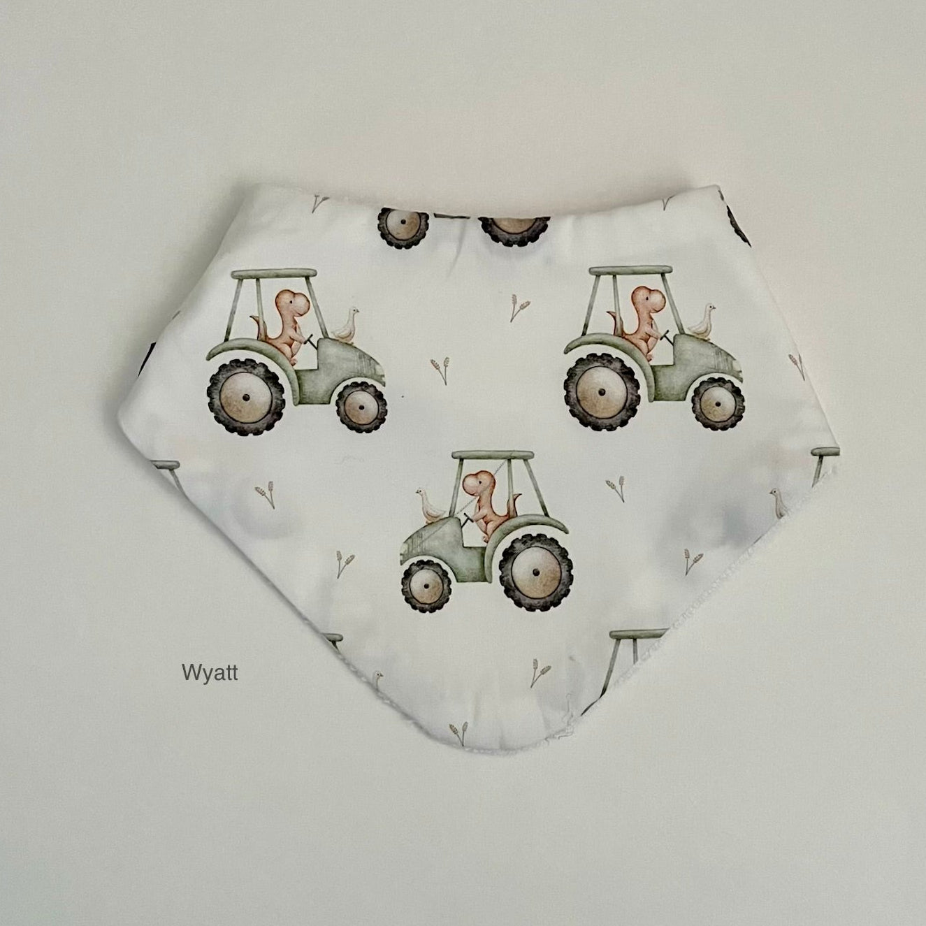 A white triangular bandana bib featuring a repeating pattern of light green tractors with cartoon figures in the driver's seat, displayed flat on a light gray background.