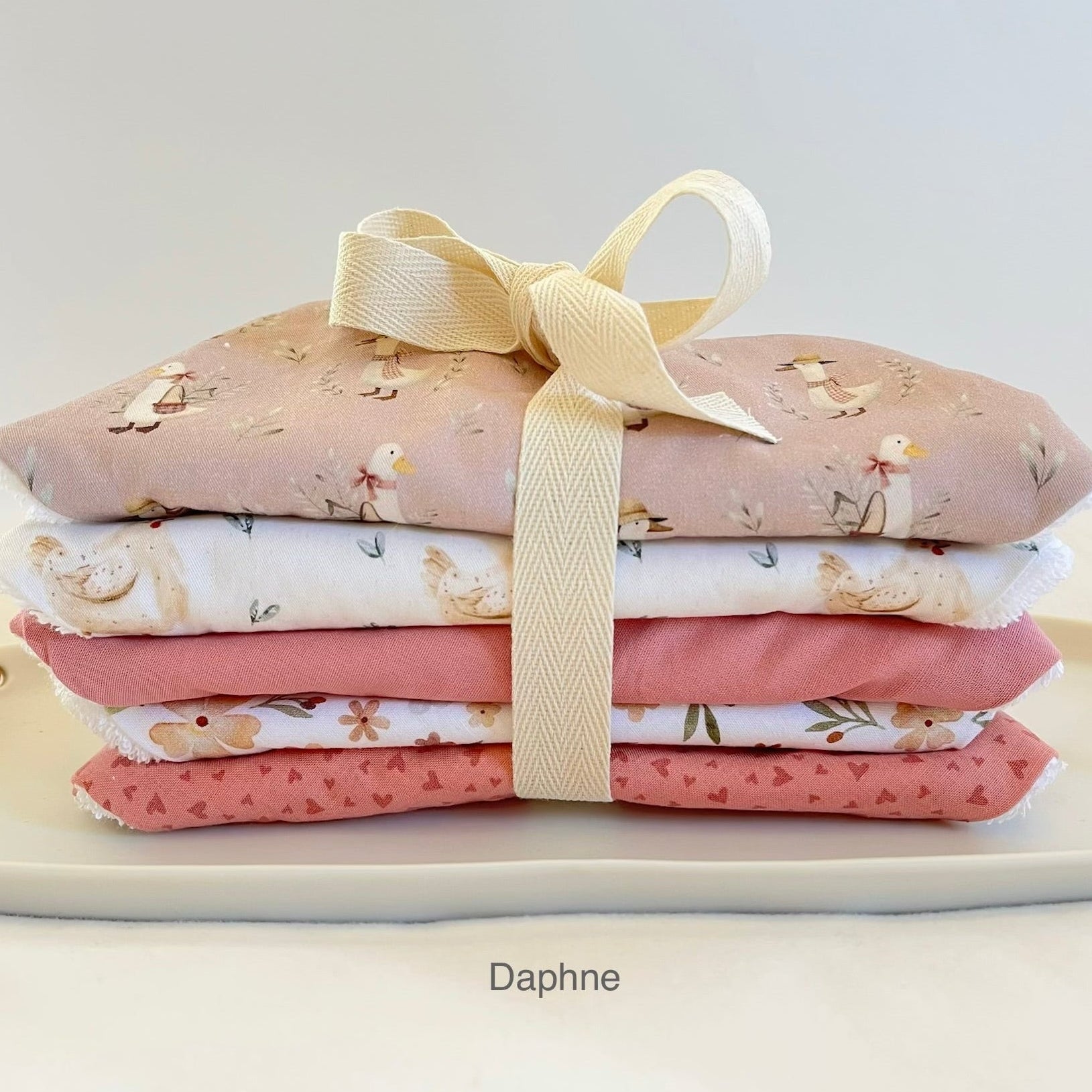 A stack of  folded fabric bibs tied with a cream ribbon, displayed on a white surface. The fabrics feature soft pastel patterns including ducks, hearts, and leaves.