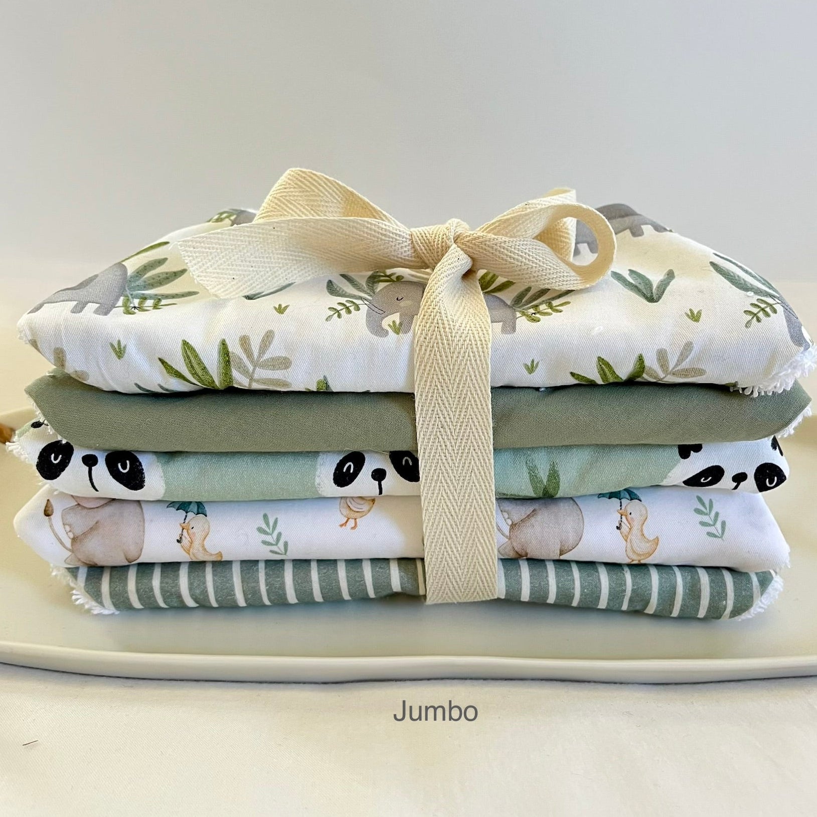 A stack of five folded baby bibs tied with a cream ribbon, featuring woodland animal prints like pandas, squirrels, and a deer on a white background, with sage green and striped patterns. The word 'Jumbo' is printed below the stack.