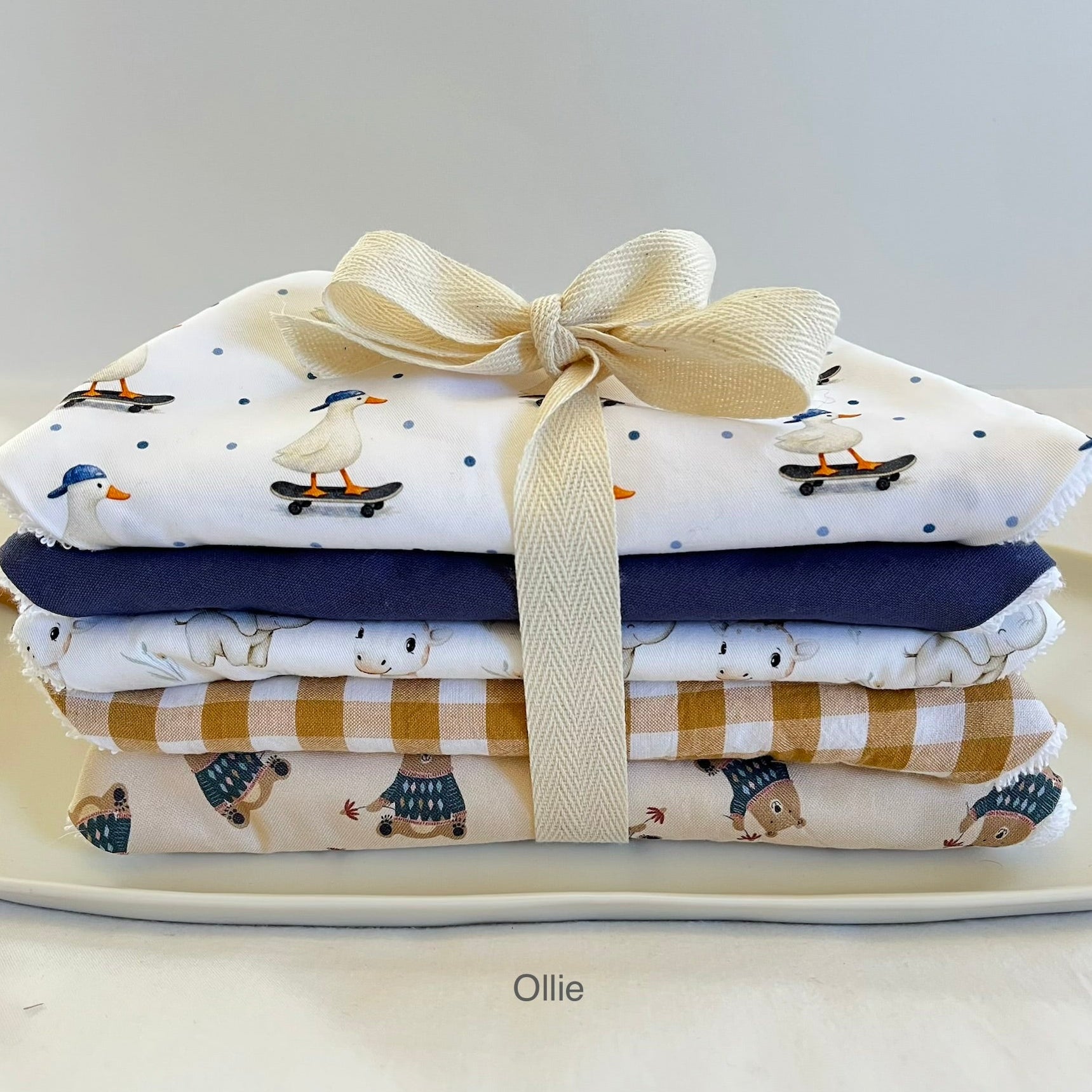 A stack of four folded baby bibs tied with a cream ribbon bow, displayed on a white plate. The bibs feature various cute prints including ducks on skateboards, polar bears, and a mustard-and-white gingham pattern.