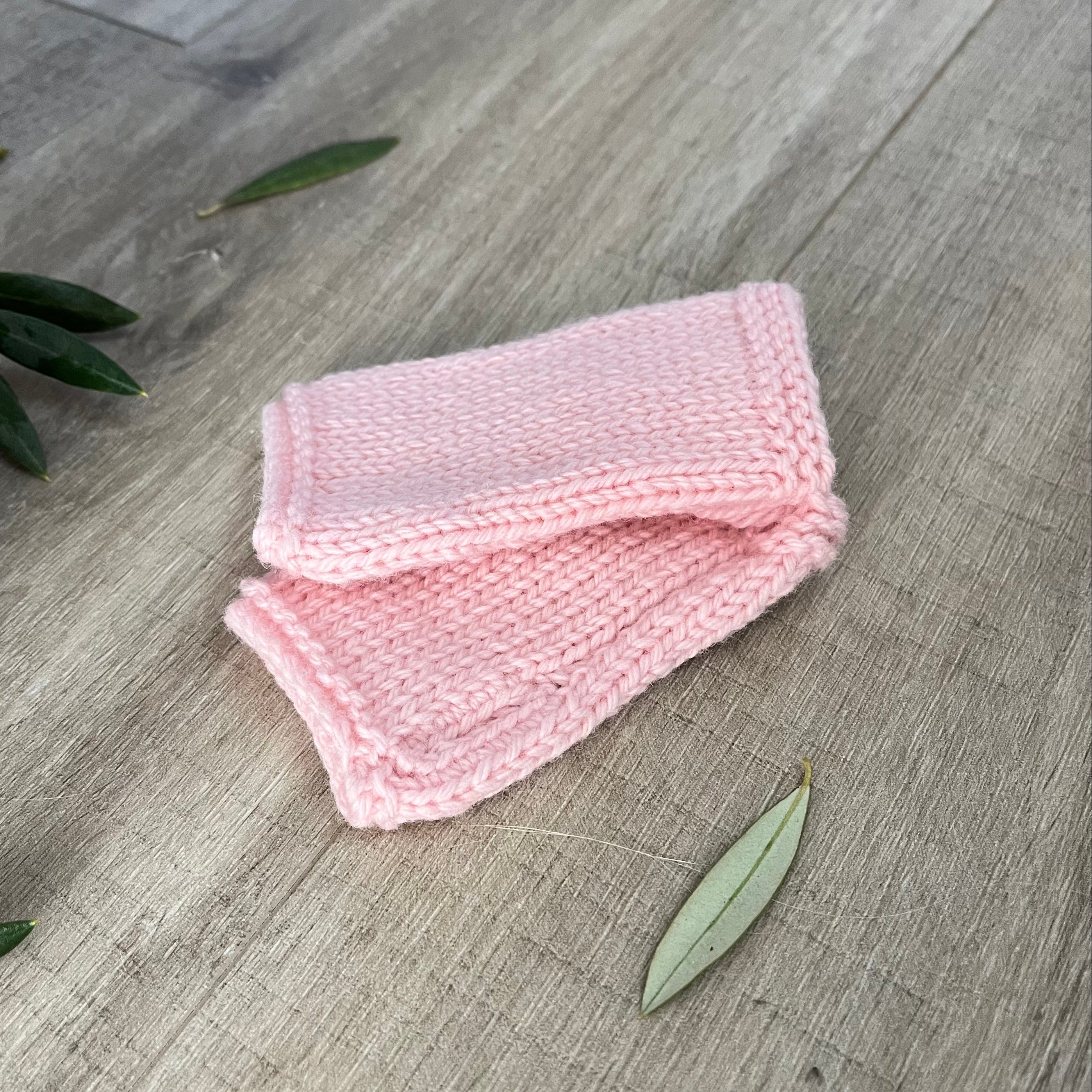 Pink knitted fingerless mittens on a wooden surface with olive leaves.