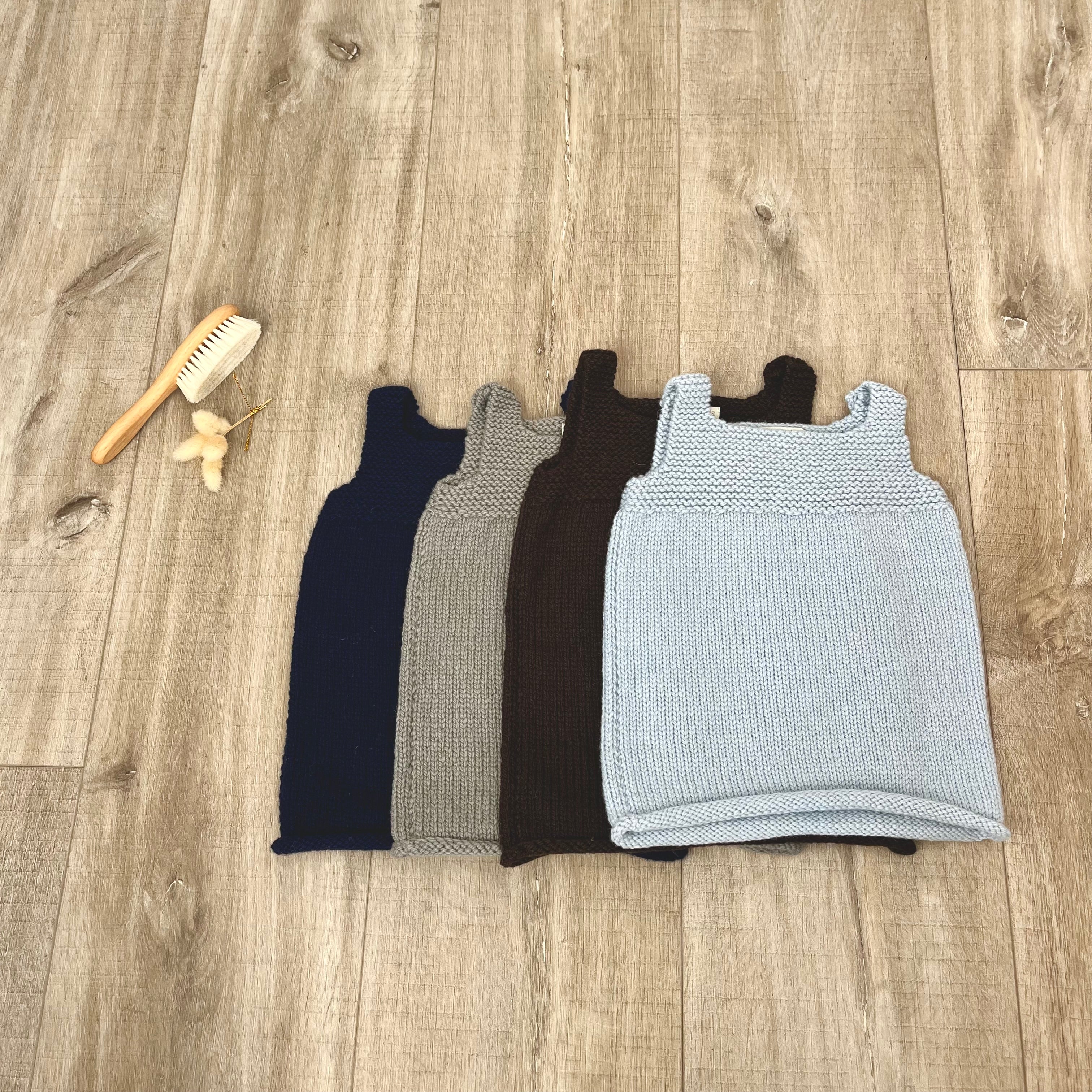 Four knitted sleeveless baby dresses in navy, gray, brown, and light blue, arranged on a wood floor next to a small wooden brush and a small beige accessory.
