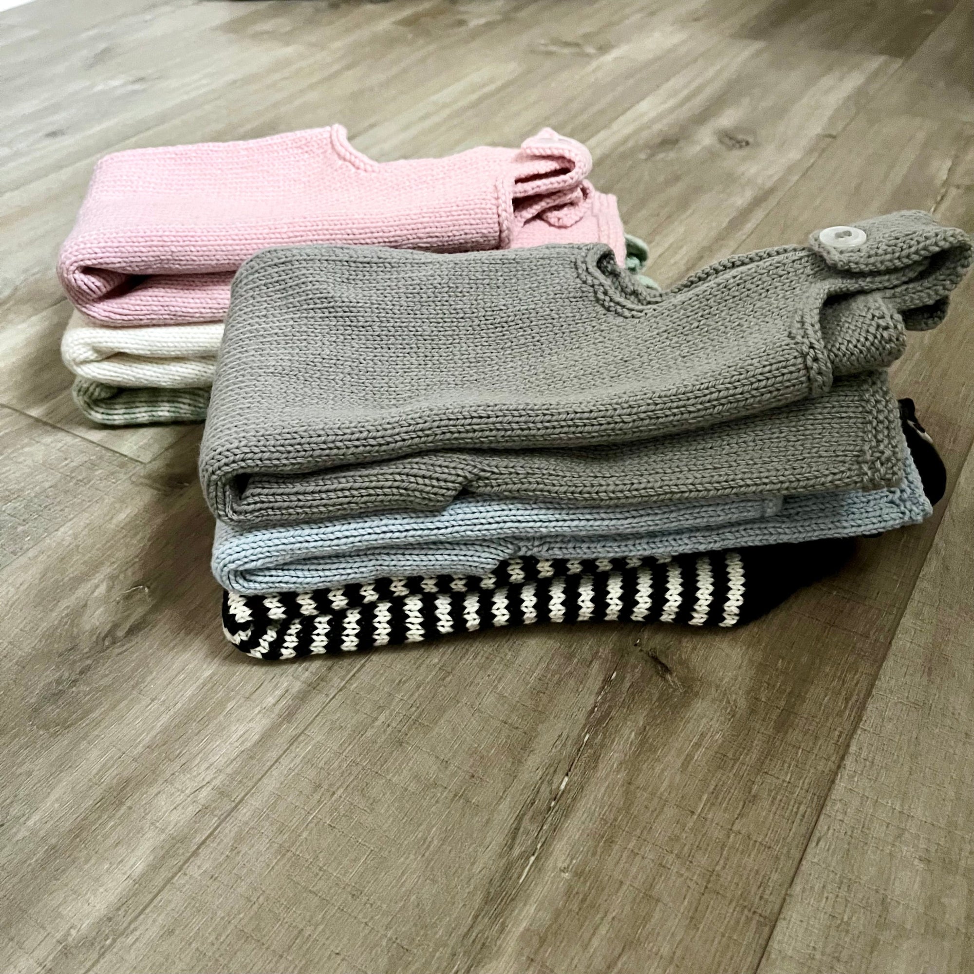 A stack of folded knit baby dungarees displayed on a light wood floor. The top item is a taupe-gray pair with a snap tab, followed by a light blue pair, a black and white striped pair, and a soft pink pair in the back.