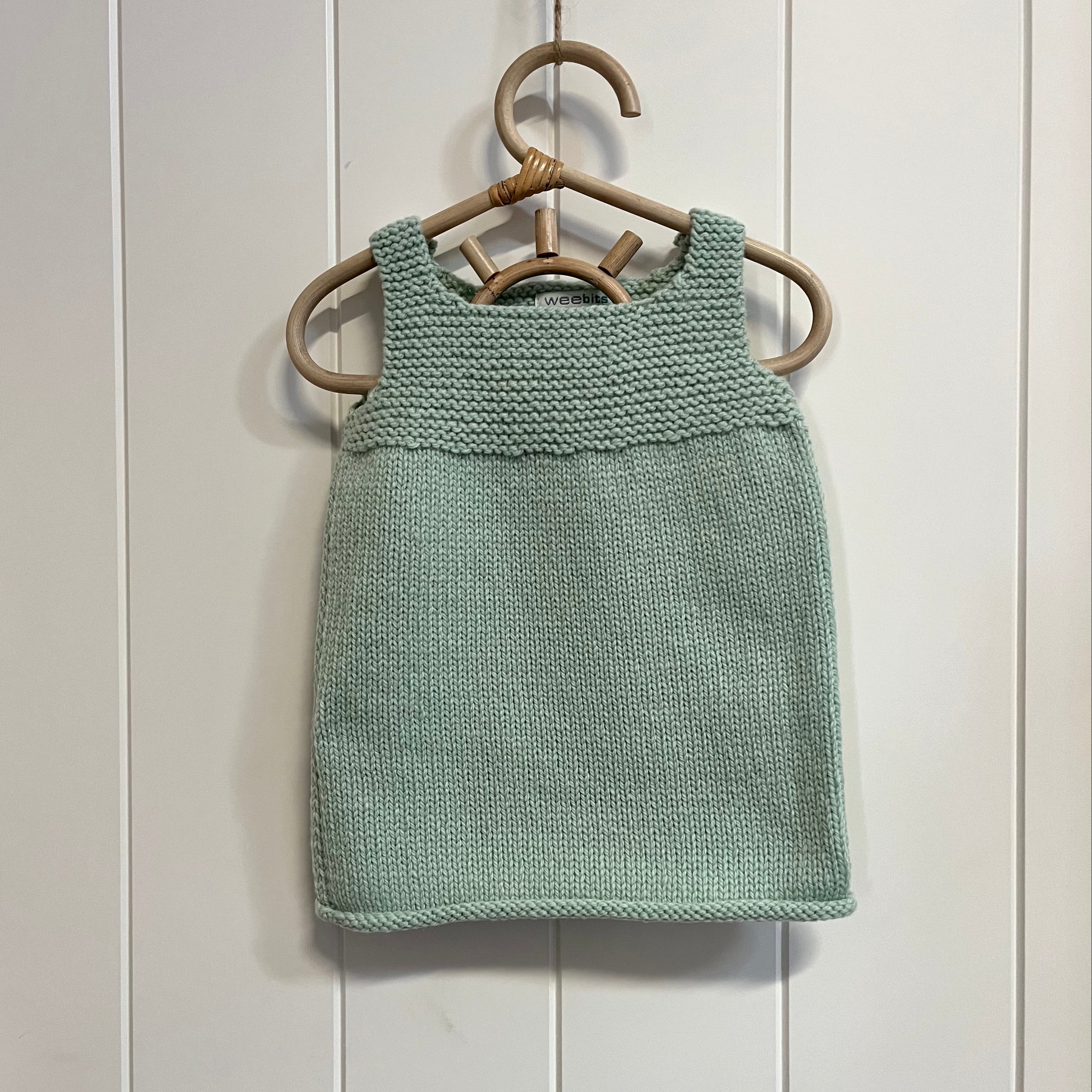 A mint hand-knit sleeveless  dress hanging on a light wood hanger against a white paneled wall.