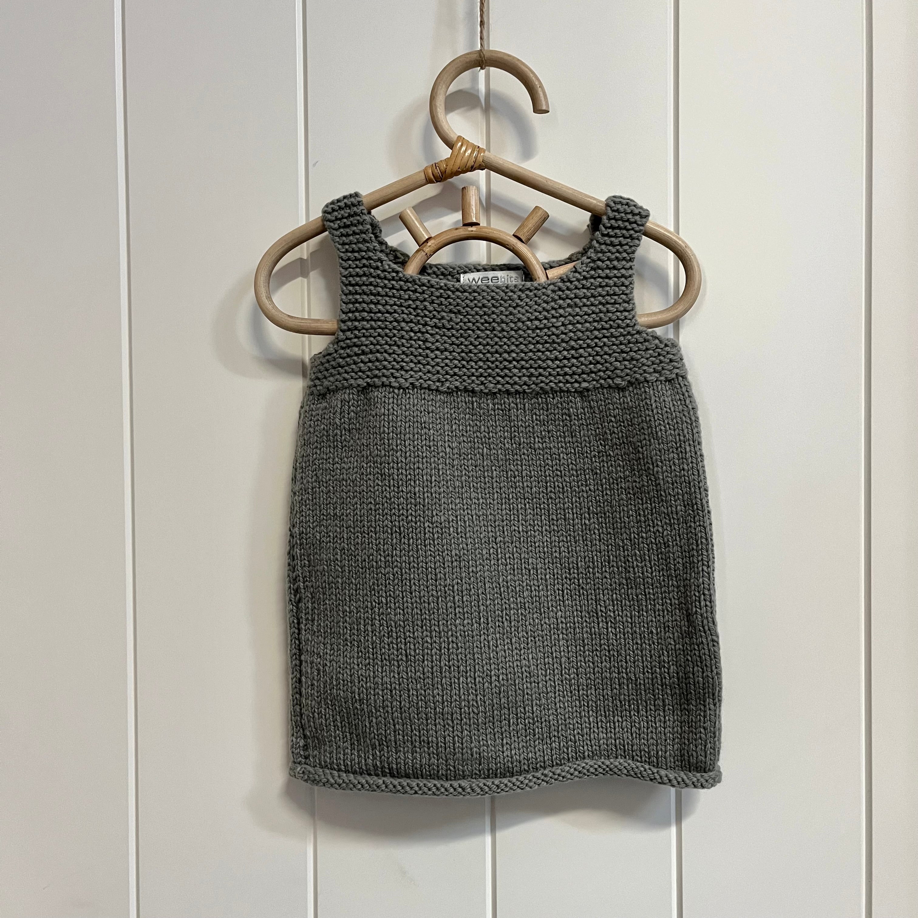 A mushroom hand-knit sleeveless  dress hanging on a light wood hanger against a white paneled wall.