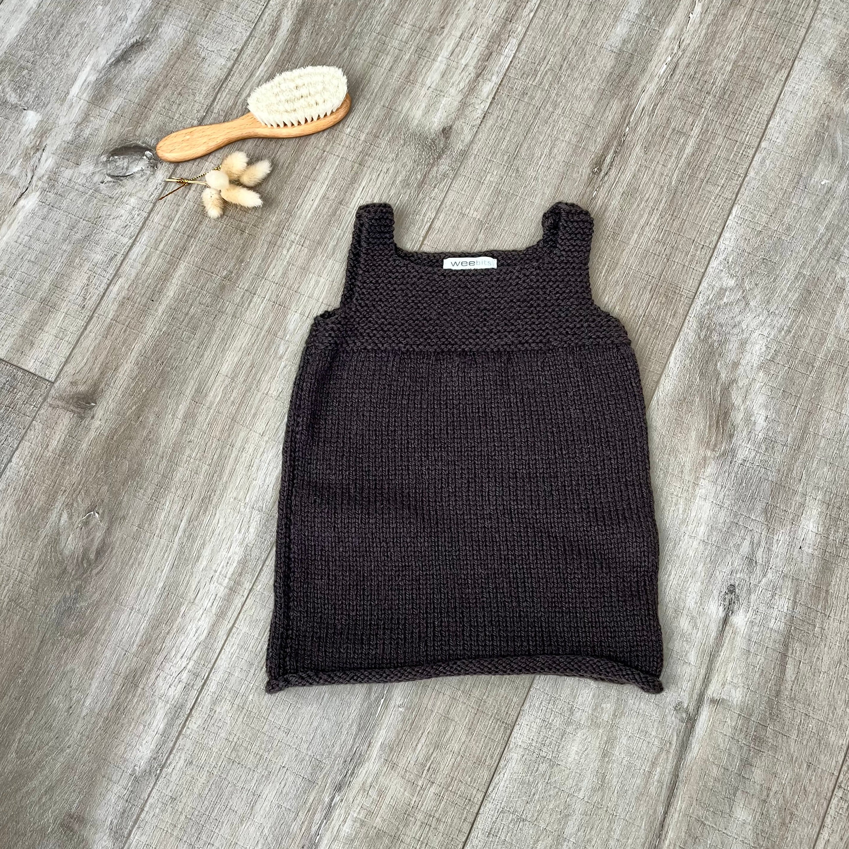 A chocolate knit sleeveless baby dress laid flat on a light brown wooden surface, styled with a small orange-handled hairbrush and dried flowers near the top left.