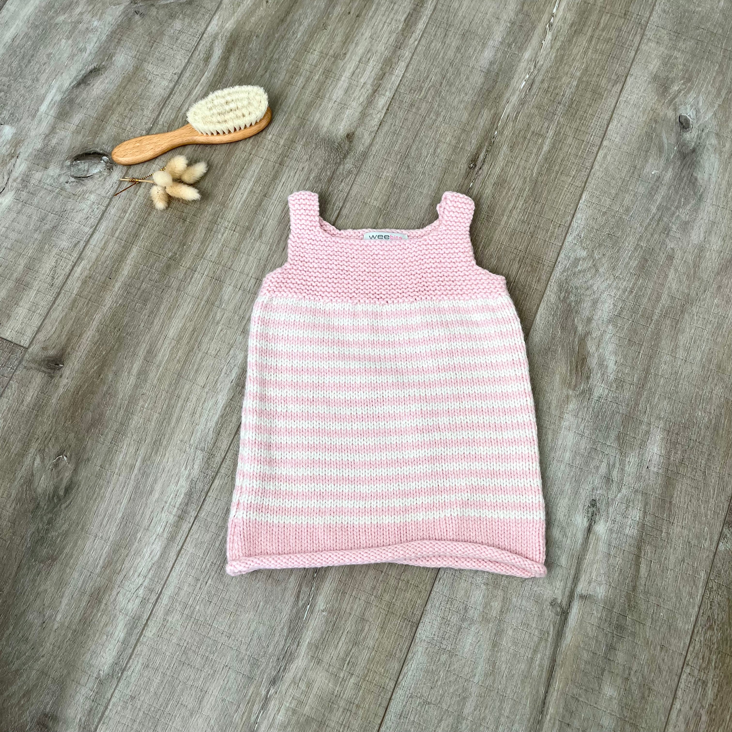A pink stripe knit sleeveless baby dress laid flat on a light brown wooden surface, styled with a small orange-handled hairbrush and dried flowers near the top left.