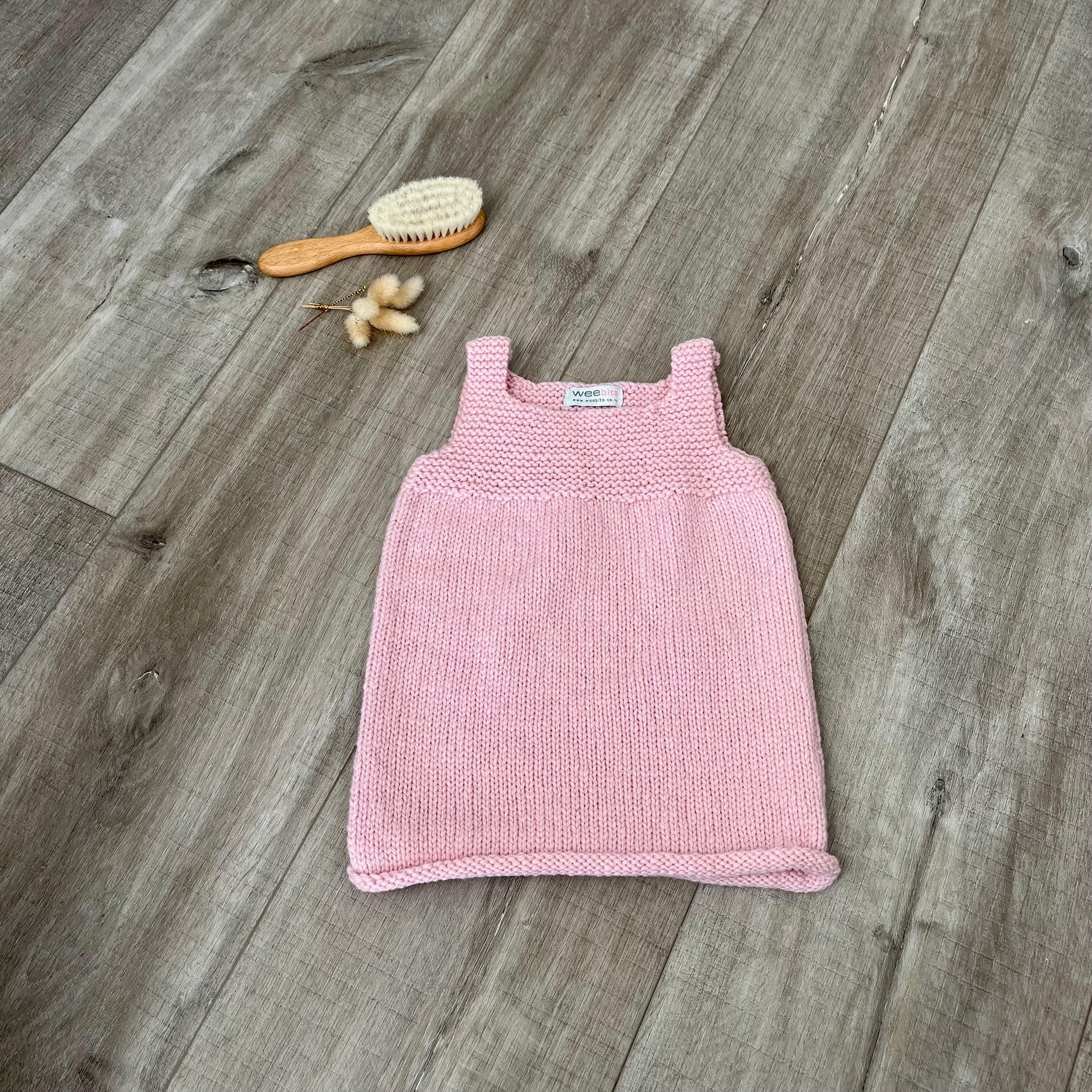 A pink knit sleeveless baby dress laid flat on a light brown wooden surface, styled with a small orange-handled hairbrush and dried flowers near the top left.