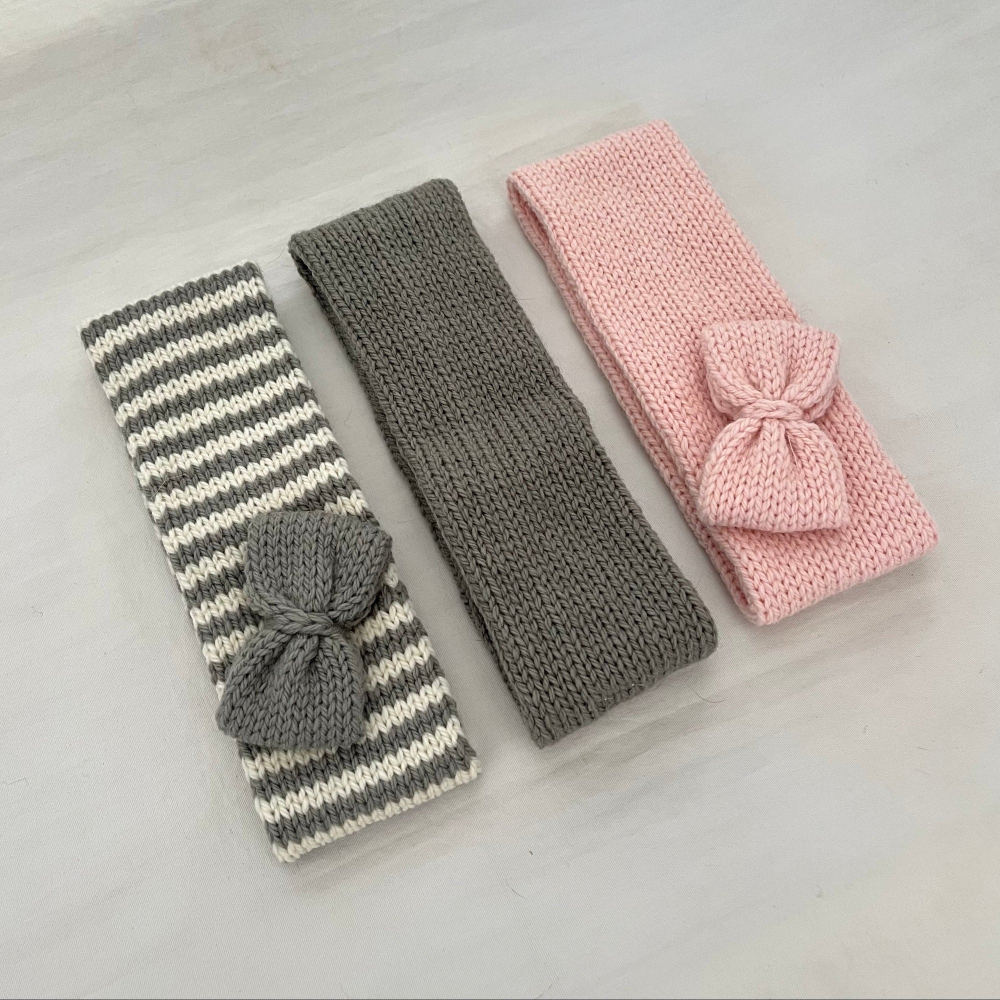 Three knitted baby headbands with bows, laid flat on a light gray surface. One headband is gray and white striped with a gray bow, one is solid gray, and one is light pink with a pink bow.
